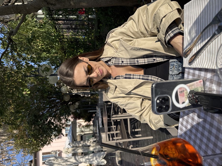 An adult female is seated at an outdoor cafe table in Sirmione, Italy, during daytime. She has long brown hair and wears dark, narrow, oval-shaped sunglasses, a light beige trench coat over an unbuttoned blue and white plaid shirt revealing a black top, and blue denim jeans. Her gaze is directed downward, focusing on a mobile phone positioned upright in front of her.
The table features a brown and white checkered tablecloth. To the left, a glass with an orange beverage, an orange slice, and a black straw is present. Centrally placed is an iPhone, in a clear case, supported by a small black tripod or stand. A white circular MagSafe-compatible accessory is attached to the phone's back, securing a pink/red banknote (with the numeral '1000' visible) underneath. To the right, a beige and white striped napkin holds a silver fork and knife.
The background includes several empty outdoor cafe tables with dark chairs and folded white umbrellas. Further back, a light pink building with dark windows is visible, partially obscured by green trees and decorative spherical outdoor lights. Natural sunlight illuminates the scene, creating distinct shadows.