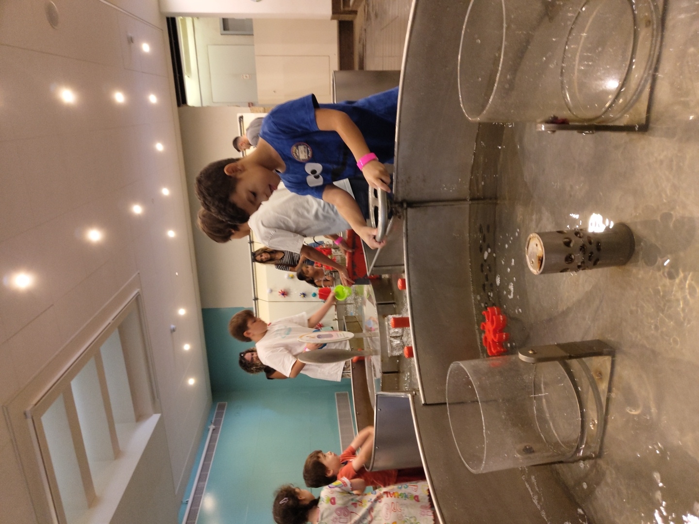 The image shows a group of children and adults at an interactive water exhibit. In the foreground, a young boy in a blue shirt is operating a steering wheel attached to a large metal water table. Another child in a white shirt is visible to his left, also engaged with the water. Several other children and adults are present in the background, some of whom are observing the exhibit. The environment appears to be an indoor recreational or educational space, with a modern ceiling and lighting. The water table itself is filled with water and contains various interactive elements, including red plastic gears. One child on the left is wearing a colorful shirt and appears to be playing with the water. The location is not identifiable from the image itself, and therefore cannot be placed in Haifa, Israel.