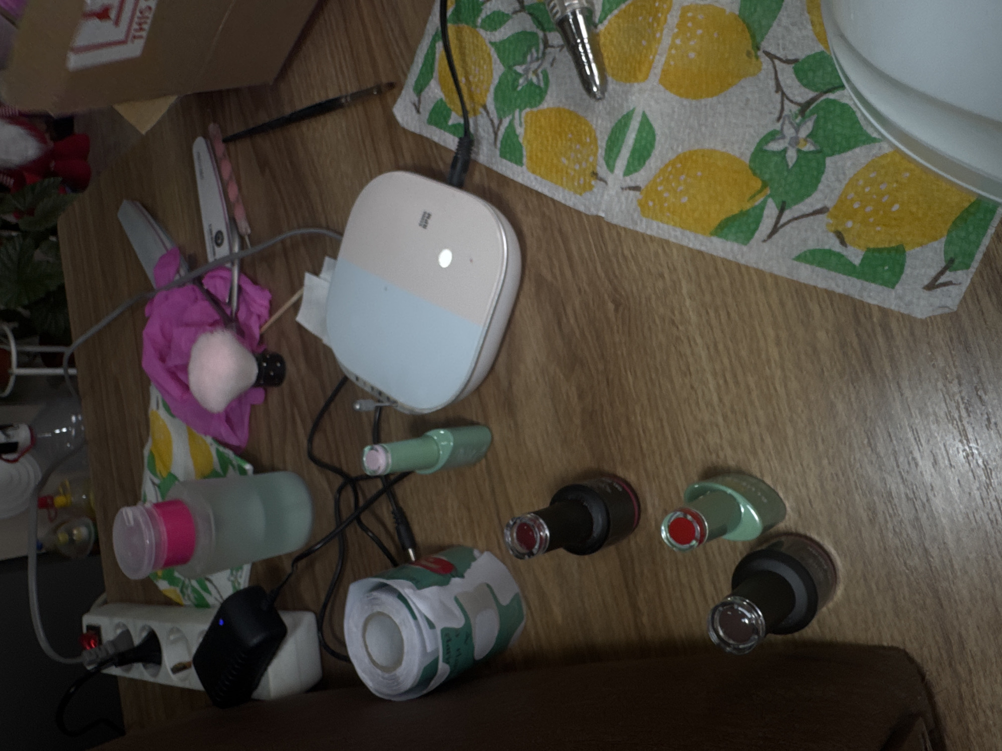 The image depicts a cluttered tabletop scene, likely in a personal space. The surface has a wood-grain pattern.
Objects present include:
* A partially visible cardboard box.
* Nail care items: nail files, a pink buffer, a bottle with a pink lid, three nail polish bottles (red, green, and brown), a green cuticle pusher.
* A white and light blue electronic device.
* A lemon-patterned towel.
* A small plant.
* A power strip.
* A roll of patterned tape.
There is no indication of the location being in Lehchevo, Bulgaria, nor any indication of people, subjects, or actions. The scene appears static and focused on nail care equipment.
