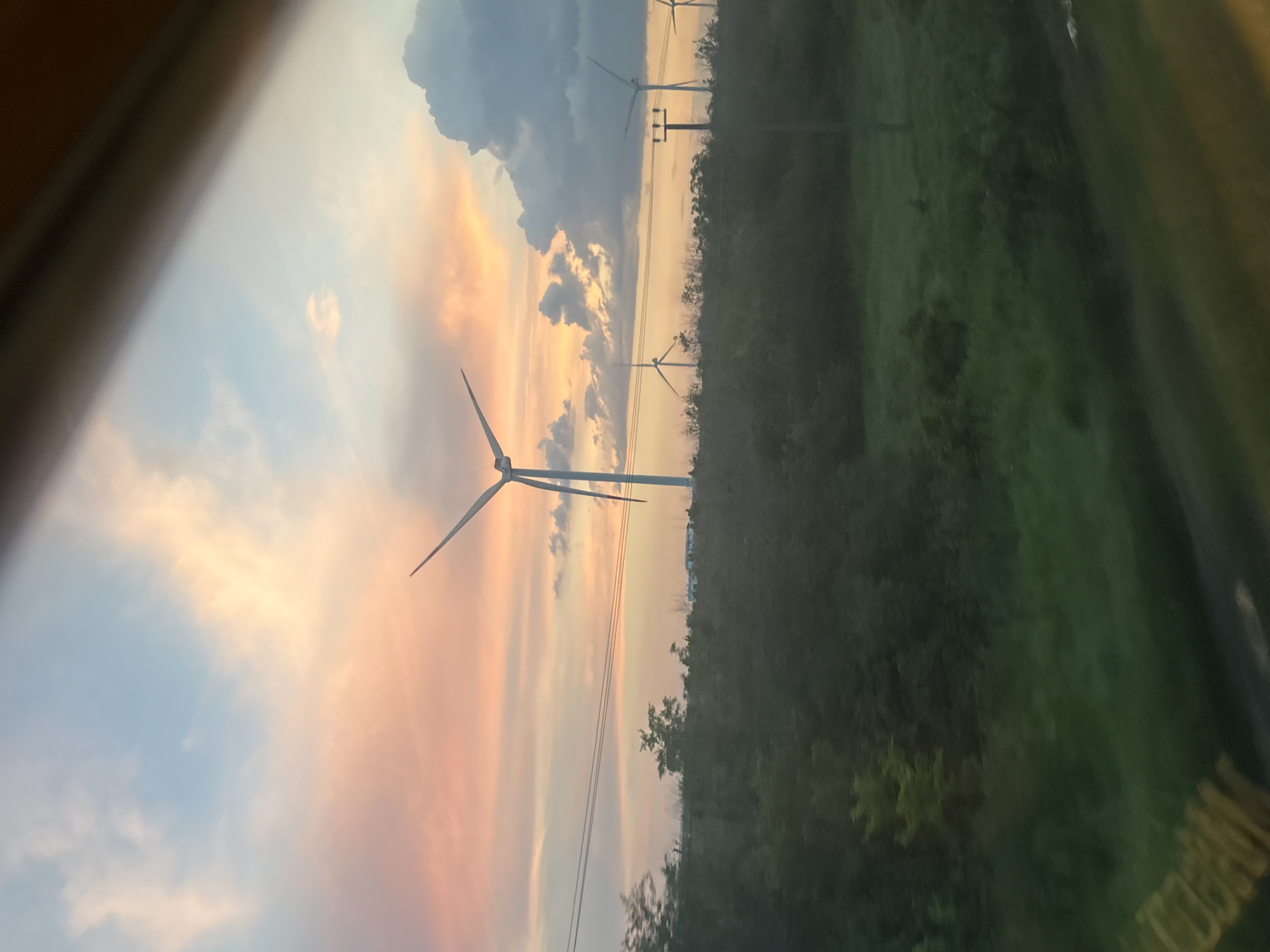The image shows a rural landscape at sunset, with a sky filled with pink and orange clouds. Several wind turbines are visible in the distance, along with power lines and a line of trees. The foreground is a dark green field with some lighter green patches. The scene is viewed through what appears to be a window, as indicated by the blurred brown and dark gray frame at the top of the image. The location is specified as Verkhnegoveinyi, Russia.