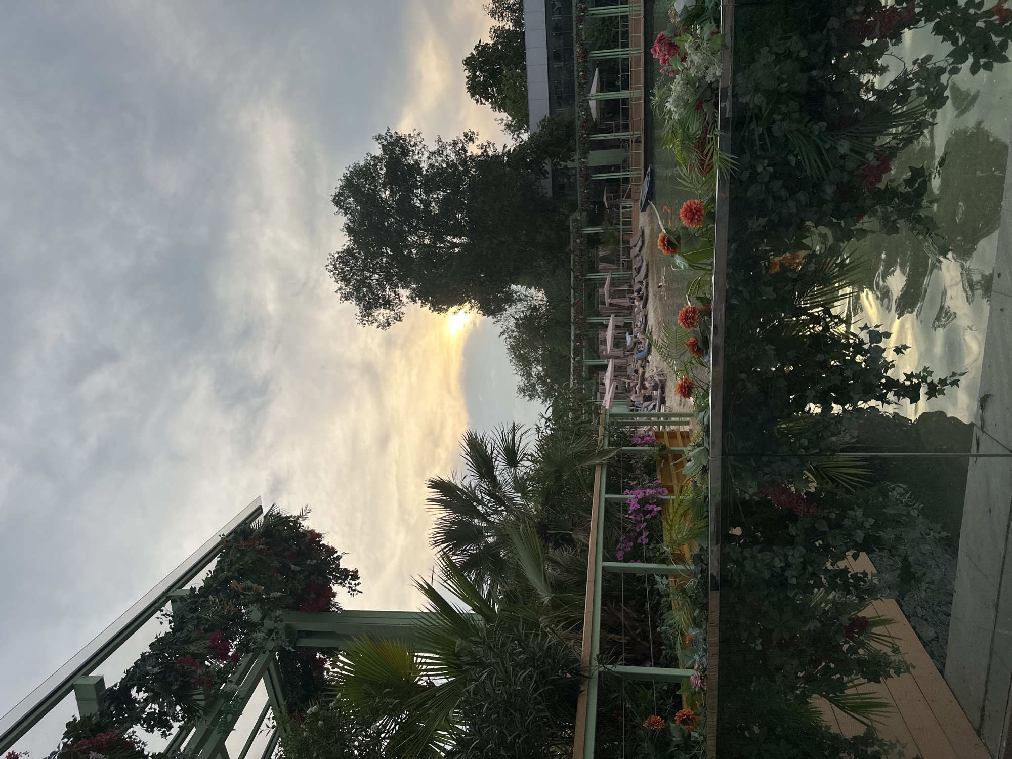 The image depicts an outdoor scene near a body of water, possibly a pool or a lagoon, with lush vegetation and floral arrangements. In the foreground, a reflective surface, likely water, mirrors the greenery and the sky above. A barrier, possibly a railing, separates the foreground from a lounging area with numerous deck chairs, some occupied by people. Beyond the lounging area, a structure with thatched roofs and decorative drapes suggests a resort or an entertainment venue. The sky is overcast with the sun visible as a bright, hazy orb behind the clouds, indicating either sunrise or sunset. The overall ambiance suggests a tranquil and resort-like setting.