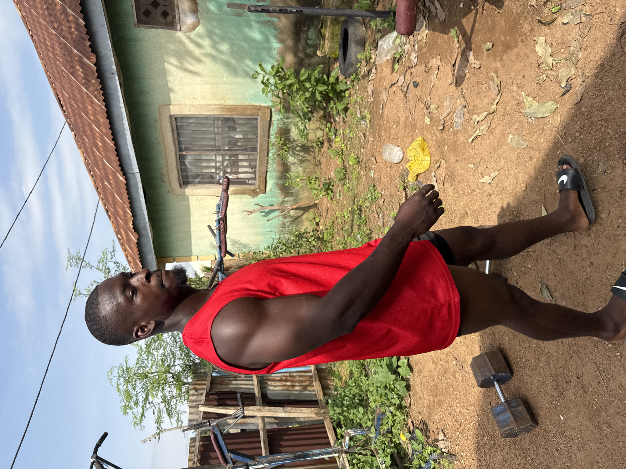 A Black man wearing a red tank top and black shorts is walking outdoors in Akwanga, Nigeria. He is on a dirt path in front of a green building with a rusted metal roof. To his left, there is a homemade weightlifting apparatus and a dumbbell lying on the ground. The area is somewhat unkempt, with scattered leaves, plastic bags, and a deflated tire. The sky is clear and blue.