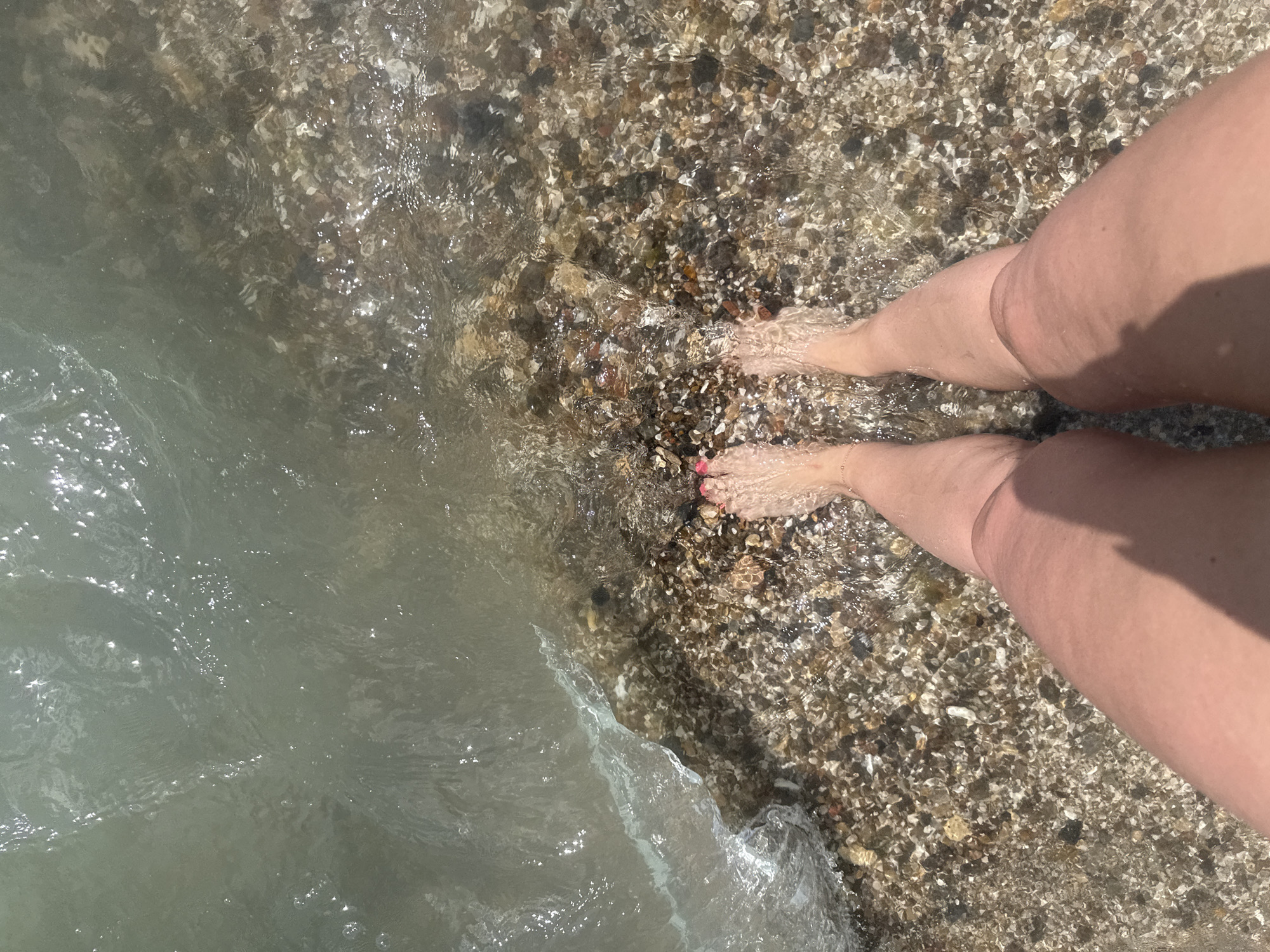A high-angle shot captures a person's legs and feet partially submerged in clear, shallow water at the edge of a pebbled shore. The water, reflecting sunlight, creates shimmering patterns on the surface and reveals the colorful pebbles beneath. The person's feet are bare, with toenails painted pink, and a delicate ankle bracelet is visible on the left ankle. The pebbled shore transitions from the water's edge to dry land. The scene is set on a beach in Odesa, Ukraine, likely during daylight hours.