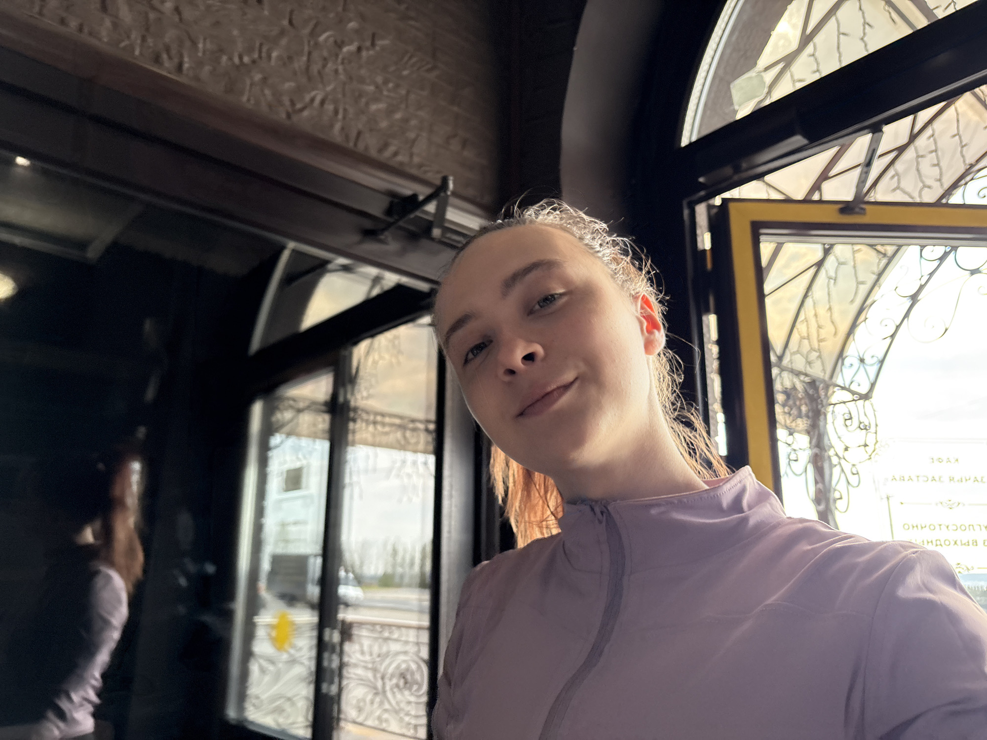 A young woman poses for a selfie indoors, with the scene framed by dark architectural elements, possibly part of a restaurant or cafe interior. Through the ornate window behind her and to her right, one can see daylight and architectural details reminiscent of Verkh-Sosnova, Russia, with reversed text visible indicating it may be signage from a building across the street. A second window or entrance to the left, with a woman standing in front of it, providing a glimpse of outdoor conditions. The young woman wears a light purple top with a high collar, and the lighting suggests it might be a bright, overcast day outside.