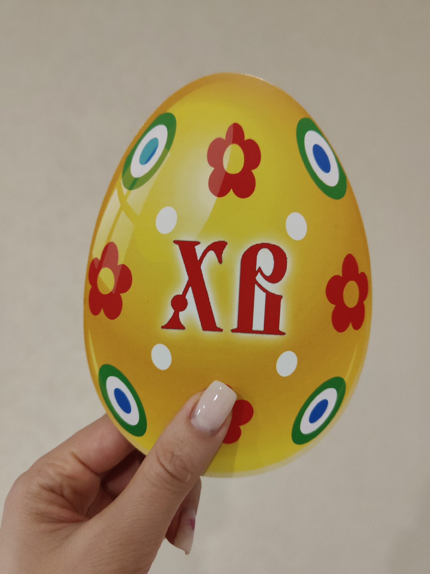 A hand holds a yellow Easter egg decoration with red flowers, white dots, and blue/white/green concentric circles on a beige background. The egg also has the red Greek letters "ΧΡ" on it. The specific location in the city of Unknown, Unknown, is not discernible from the image.