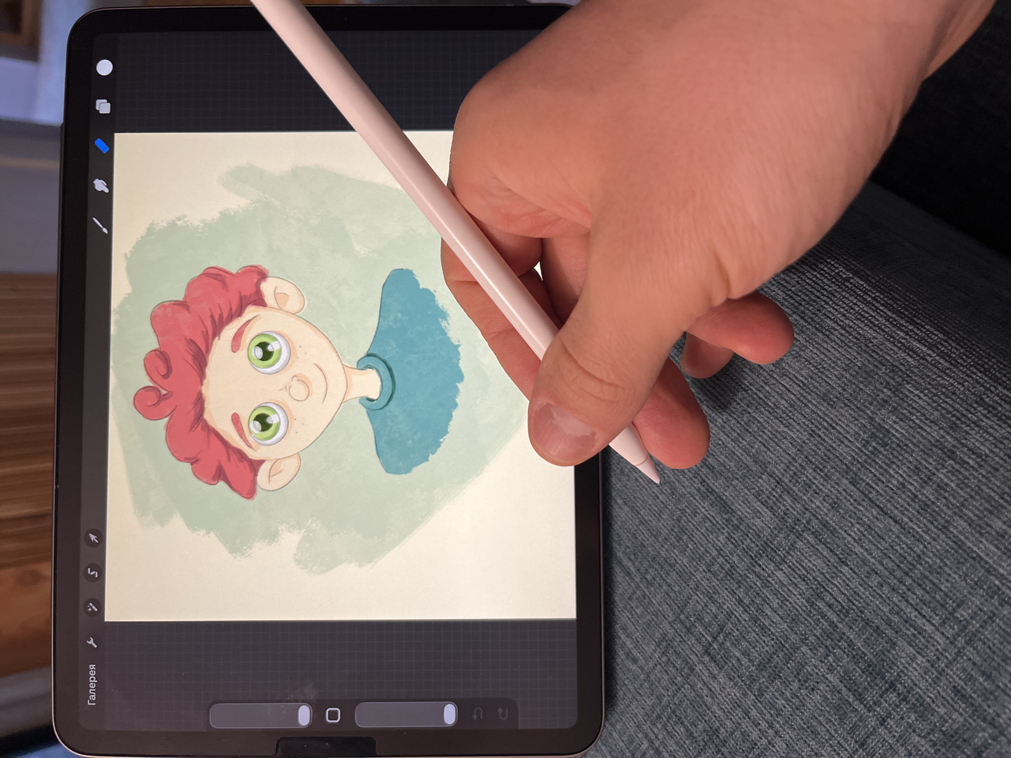 A hand holds a pink stylus over a tablet displaying a digital illustration of a cartoon character with red curly hair, green eyes, and a blue shirt against a light green textured background. The tablet's user interface is visible on the left side. The background appears to be a wooden surface, and the tablet is resting on a textured blue fabric. There is no indication of the location being in Tashkent, Uzbekistan.
