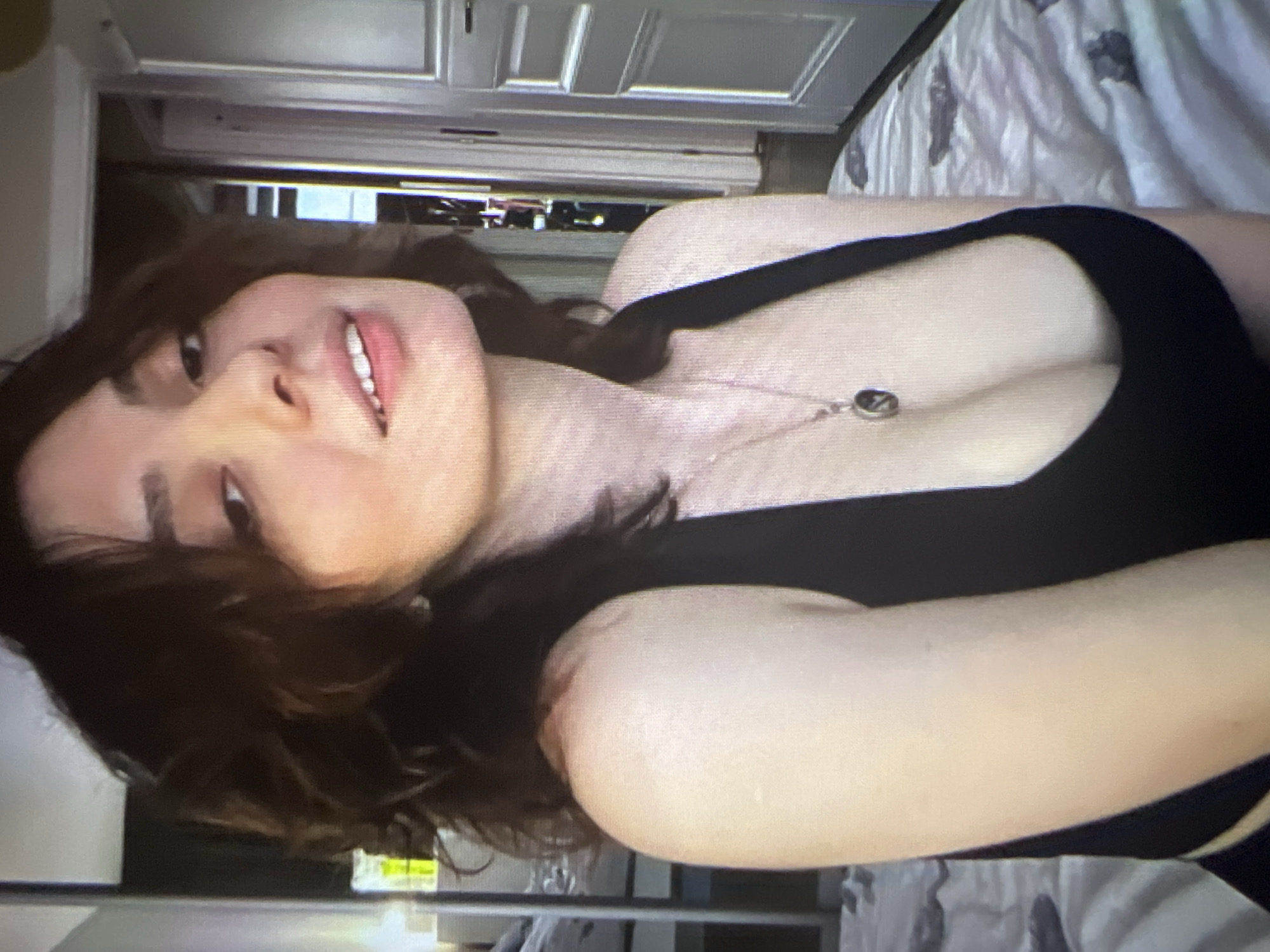 The image shows a young person with dark, wavy hair, tilted to the right. They have fair skin and are wearing a black tank top. A delicate necklace with a small, dark pendant hangs around their neck. The person's face is partially obscured by their hair, but their eyes are open, and their lips are slightly parted, revealing teeth.
The background is out of focus but appears to be an indoor setting, possibly a bedroom. A white duvet with a dark pattern is visible behind the person. A doorway or closet with white doors is also discernible. The lighting suggests daytime. There is no visible text in the image. The overall mood is casual and relaxed.