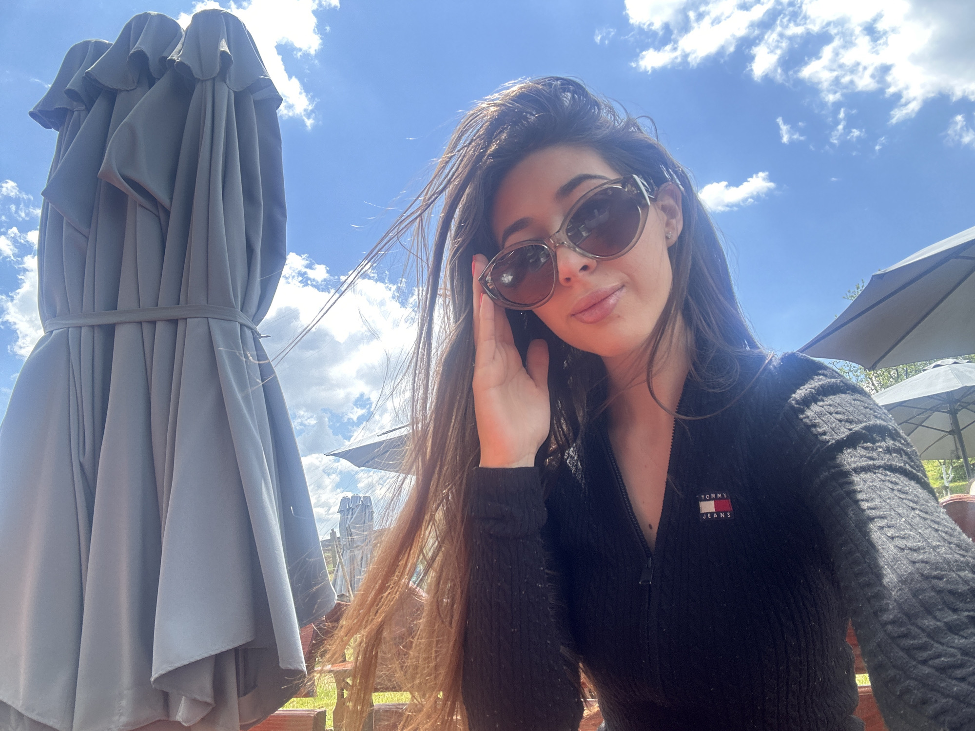 A woman poses for a selfie outdoors on a sunny day with a blue sky and scattered clouds. She is wearing dark-tinted sunglasses and a zipped-up black sweater with a "Tommy Jeans" patch on the upper left side. She has long, brown hair and holds one hand near her face. Behind her, there is a closed gray outdoor umbrella, part of a wooden picnic table, and another open outdoor umbrella in the background. The location is suspected to be in the city of Meshtitsa, Bulgaria.