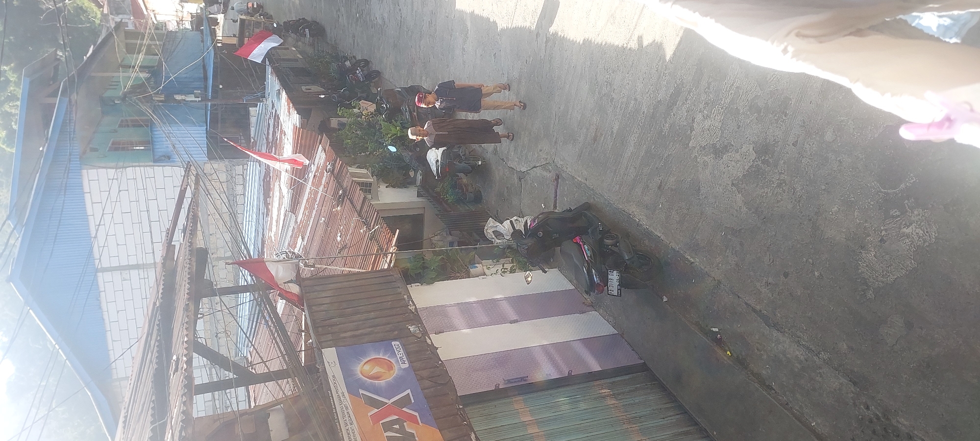 This is a high-angle, full-shot, outdoor, daytime image of a narrow, paved alleyway in a densely populated urban area. The alley is flanked by multi-story buildings with corrugated metal roofs, some appearing weathered. Indonesian flags, red and white, are visible on poles and attached to structures. Two young males, likely children or teenagers, are walking away from the camera down the alley. The male on the left wears a dark brown robe and a white skullcap. The male on the right wears a dark blue shirt, tan pants, and a red skullcap. Several motorcycles are parked along the sides of the alley. One dark-colored scooter with license plate "PA 3119 DA" is prominently parked on the left. The buildings show signs of varied construction, with some sections appearing to be made of concrete blocks and others of wood and metal sheeting. The overall impression is one of a modest, possibly older, residential or commercial area. The lighting suggests it is a sunny day, casting strong shadows. The area appears to be on a slope, given the angle of the alleyway. The environment is characterized by a mix of residential structures, commercial signage (one prominent sign reads "MAX" with an orange circular logo), and the general urban clutter of parked vehicles and exposed electrical wiring.