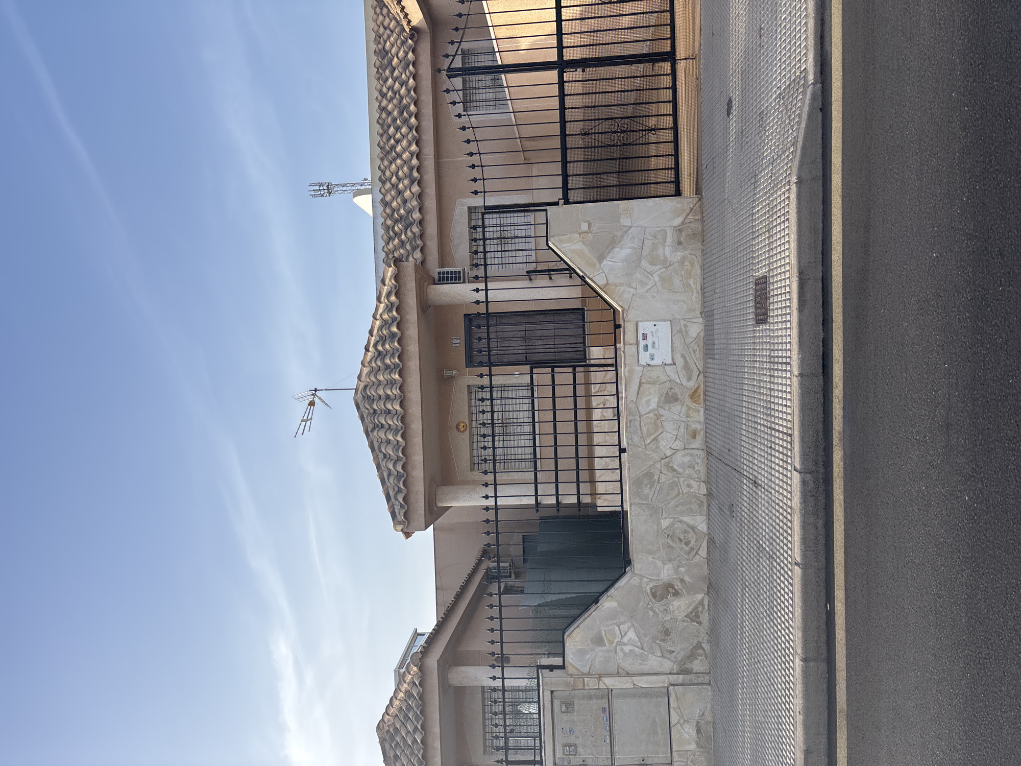 The image shows a street view of a residential area in Orihuela, Spain, under a clear blue sky with some wispy clouds. In the foreground, a dark asphalt road with a yellow line is visible, bordered by a concrete curb and a textured grey sidewalk. Behind the sidewalk, a stone-clad wall separates the property from the street.
Dominating the mid-ground are two single-story houses with beige tiled roofs and light beige walls. Both houses have black wrought-iron fences and gates. The house on the left has a dark green gate and a light grey electrical box mounted on the stone wall. The house on the right features a more ornate black gate with decorative scrollwork. Both houses have windows with black security bars or shutters. An air conditioning unit is visible on the wall of the right house.
In the background, various antennas and communication towers are visible against the sky, suggesting a well-connected urban or suburban environment. The overall scene depicts a quiet, sunny day in a Spanish residential neighborhood.