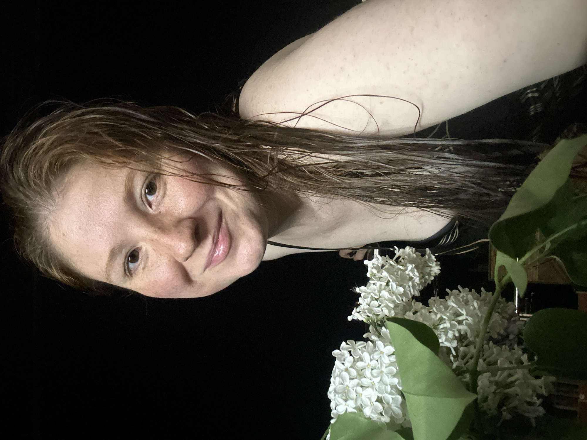 A person with wet, reddish-brown hair is positioned to the left of a bouquet of white lilacs with green leaves, set against a dark background. The person is smiling, with visible freckles and light skin tone. The person appears to be wearing a black top with thin straps. The image has a slightly dark and low-lit quality. The city of Nizhny Novgorod, Russia is not discernible in the image.