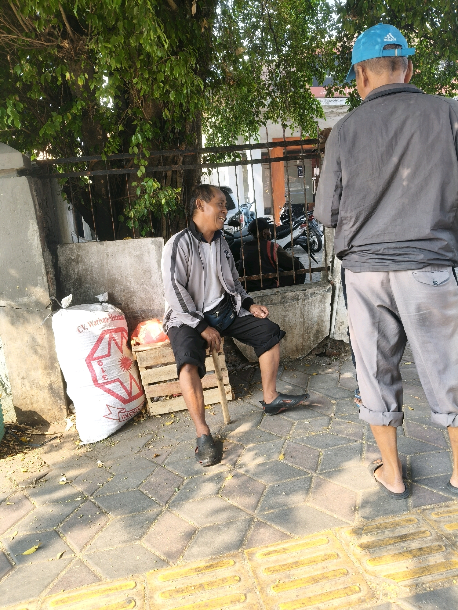 The image depicts an outdoor scene, likely on a sidewalk in South Tangerang, Indonesia. In the foreground, a man is seated on a wooden crate, wearing a grey and white jacket, black shorts, and black sandals. He is holding a wooden stick and appears to be smiling and looking to his left. To his left is a large white sack with red lettering. Behind him, another man wearing a grey jacket and grey pants stands facing away from the camera. He is wearing a blue baseball cap. In the background, there is a metal fence with trees and foliage behind it. Further in the background, motorcycles are visible. The ground is paved with interlocking bricks and features tactile paving for the visually impaired. The lighting suggests it is daytime, with sunlight casting shadows.
