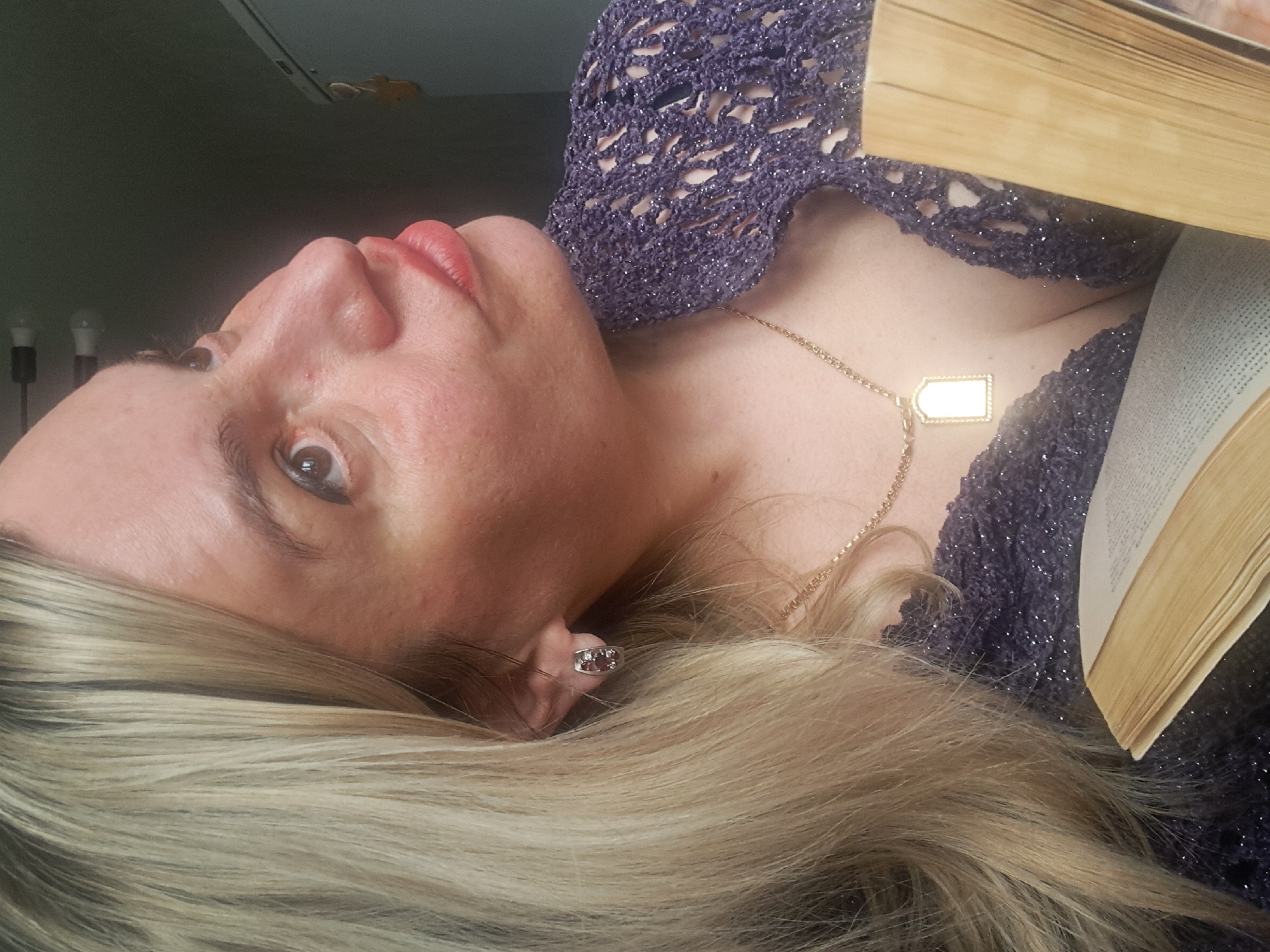 The image is a close-up, rotated portrait of a woman indoors, appearing to be reading or pausing from reading a book. She has long, straight blonde hair, fair skin, and light brown eyes, with visible dark eyeliner and light pink lipstick on her slightly parted lips.
The woman is wearing a dark purple, open-knit or crocheted top with sparkling threads. Around her neck is a thin gold chain necklace featuring a bright, rectangular golden pendant that reflects light, making any details or text on it illegible. A silver-toned earring with several small stones is visible in her right ear, partially obscured by her hair.
An open book with numerous light-colored pages and a light brown spine or cover is positioned near her, partially covering her chest and shoulder. Her gaze is directed upwards and towards the viewer, with a neutral or contemplative expression.
The setting is indoors, likely a home environment. In the background, partially visible, are two light bulbs or lamps and a portion of a ceiling or light fixture. A small, decorative object, possibly a dried leaf or star, is also attached to the ceiling or wall. No discernible text is visible on the book pages. The provided location context for this image is Elektrostal, Russia.