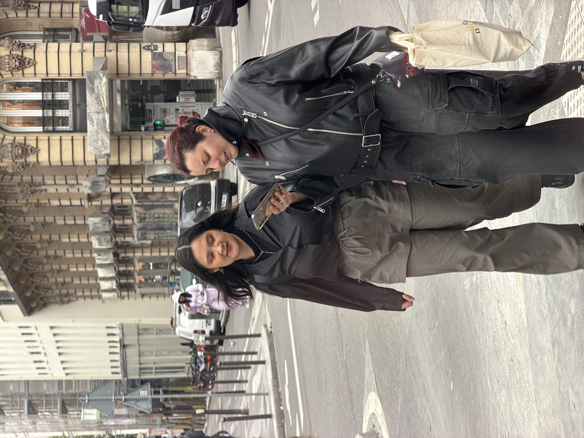 The image shows two women standing on a city street in what appears to be Paris, France. The photo is taken from a slightly high angle, looking down.
The woman in the foreground (left when upright) has dark hair and a warm complexion. She is wearing a dark top, possibly a jacket, over a darker t-shirt, and light brown or khaki cargo pants. A delicate gold necklace is visible. She holds a smartphone with a clear case in her right hand and is smiling gently, looking towards the viewer or the other woman.
The woman in the background (right when upright) has reddish-brown hair tied back. She is dressed in a black leather jacket with multiple silver zippers and buttons, and dark wash cargo pants. A black shoulder bag with various keychains is worn across her body, and she carries a cream-colored tote bag with "13" and other text (partially obscured) printed on it. She is looking downwards with a soft expression, seemingly at the first woman or her phone.
They are standing on a paved sidewalk next to a street. In the background, ornate classical European architecture, typical of Paris, lines the street with multi-story buildings featuring decorative cornices and window frames. Cars and parked motorcycles or scooters are visible further down the street. The sky appears overcast, suggesting it is daytime with diffused natural light. Both women seem relaxed and content.