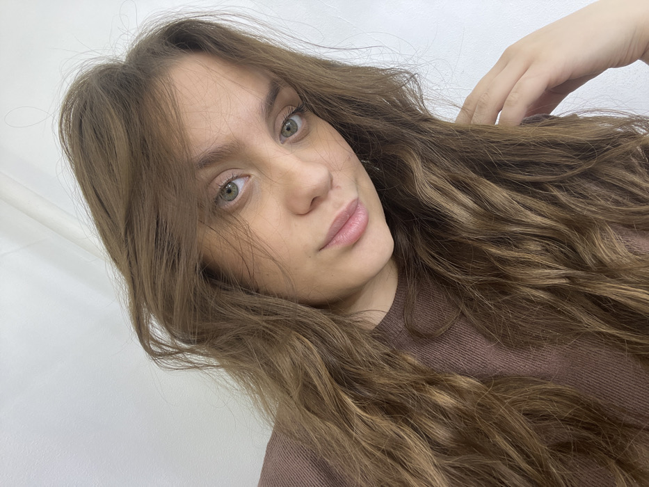 A headshot portrait features a female individual positioned against a plain white background in the city of Chaikovsky, Russia. The subject has long, wavy, light brown hair, parted slightly left, which frames her face and extends over her shoulders and chest. She has green/hazel eyes, directly engaging the viewer, and a neutral facial expression with lightly parted, natural-toned lips. Her skin tone is fair. She is wearing a brown, ribbed long-sleeved top, partially visible on the left. A portion of her left hand is present on the lower right, with fingers lightly resting near her hair or shoulder. The scene is evenly lit, indicating an indoor environment.