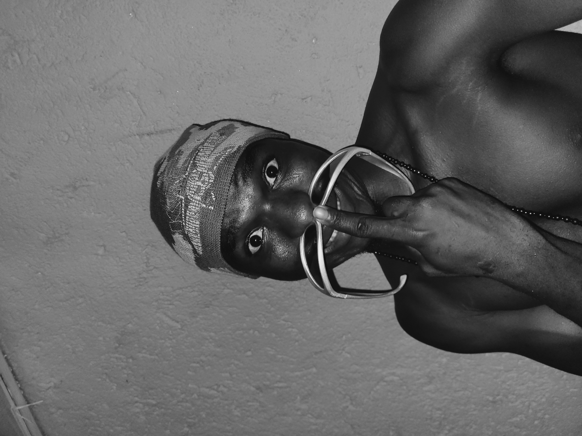 A black and white, high-angle, medium shot depicts an African male from the chest up, facing the viewer at an angle. He wears a patterned cap and a beaded necklace. He is holding a pair of rimless eyeglasses to his face with his index finger. His expression is intense, with his eyes wide open. The background consists of a textured wall. The location of the image is not evident from the visual data provided.