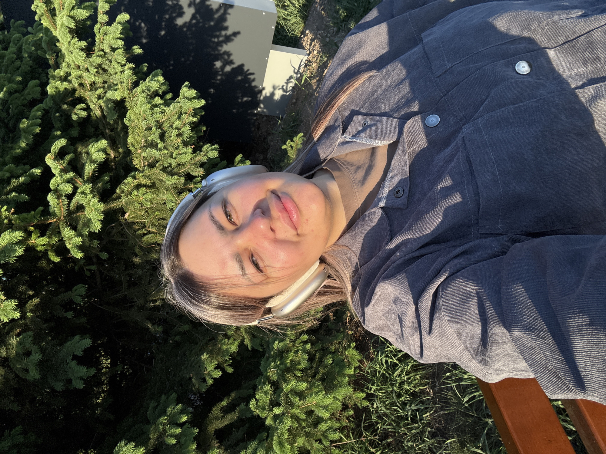 A selfie of a young woman with long blonde hair and headphones, lying on a bench in a park. The background consists of green trees and sunlight. The woman is wearing a gray jacket. The exact location within Birobidzhan, Russia cannot be determined from this image.