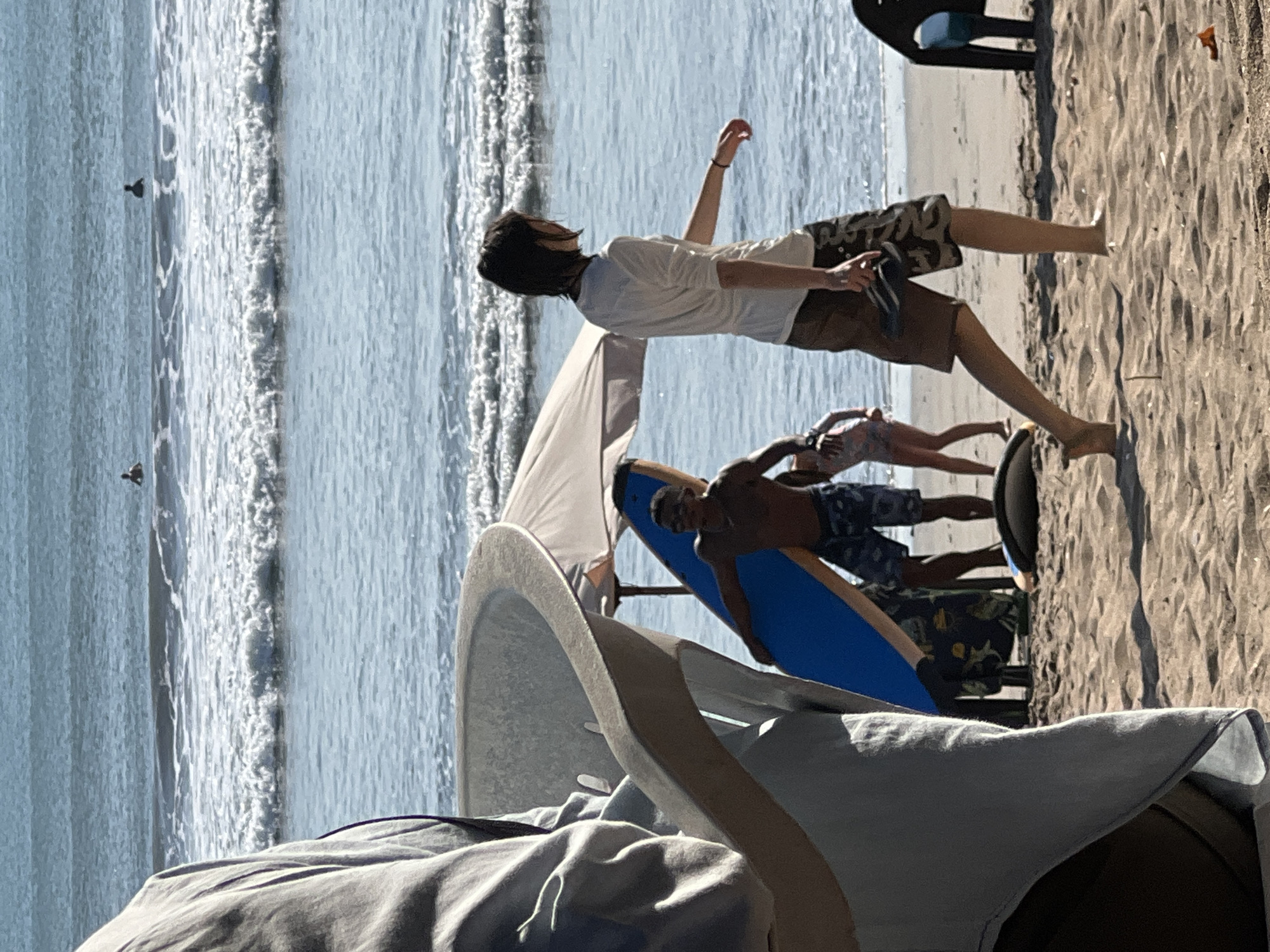 At the Kuta beach in Indonesia, the scene captures a sunny day with moderate waves. In the foreground, partially obscured by a light-colored fabric, is a dark, curved object, possibly a beach chair or seating. To the right of this, a person with dark hair and a white long-sleeved shirt is walking away from the camera, carrying a pair of flip-flops. They are wearing patterned shorts. Behind them, a shirtless person is holding a bright blue surfboard and wearing patterned shorts. A small child in a floral swimsuit is visible nearby. A beach umbrella provides shade. In the background, several surfers are in the water, waiting for waves. The sand appears dry and there is a clear view of the ocean.