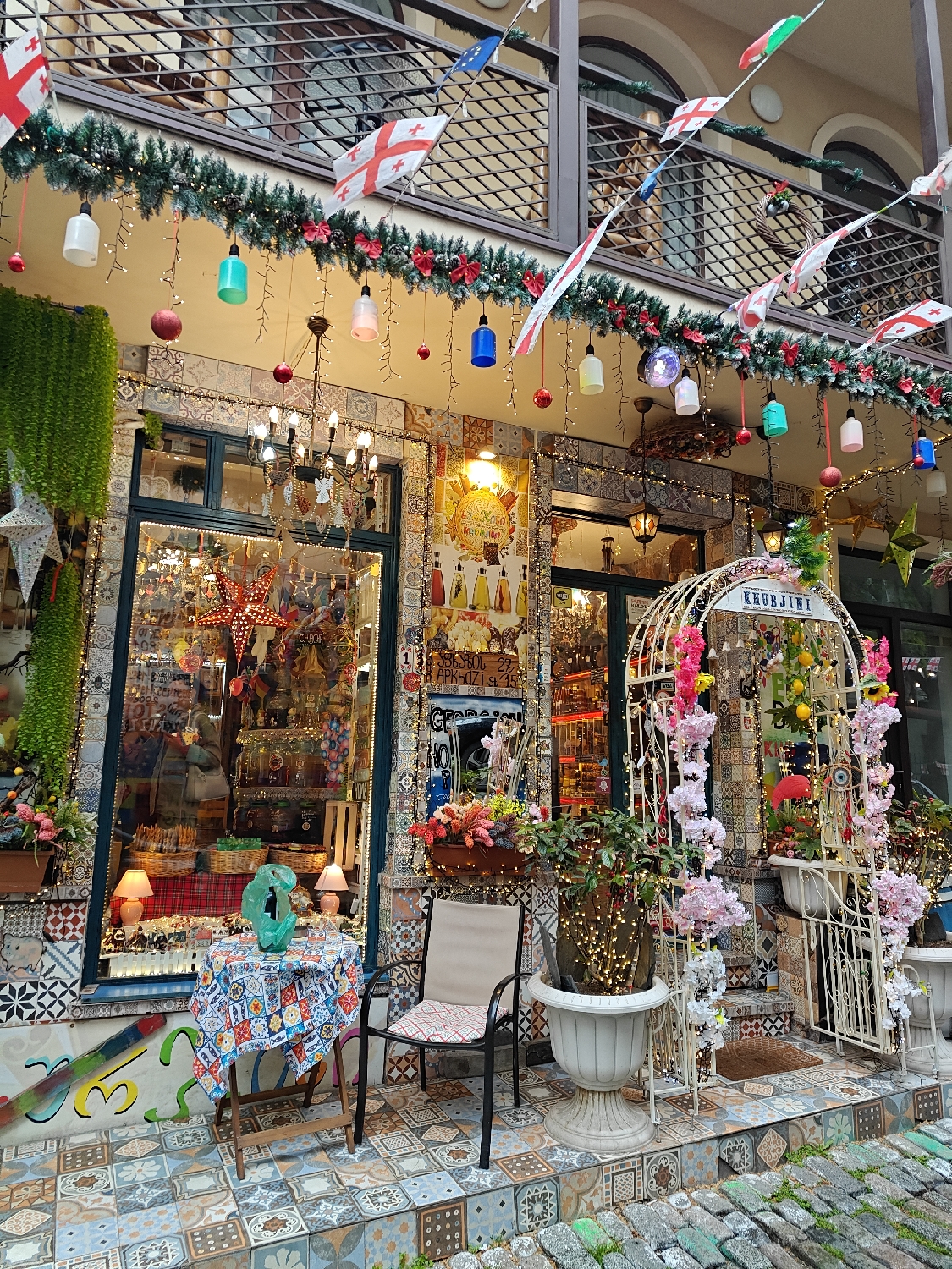 This image captures the vibrant and intricately decorated facade of a shop or cafe in Tbilisi, Georgia. The setting is an outdoor, likely pedestrian, area, with cobblestone paving in the foreground. The time of day appears to be either an overcast daylight hour or early evening, as numerous artificial lights are illuminated, creating a warm and inviting glow.
The building's exterior is a striking mosaic of colorful patterned tiles, covering the walls around the windows and doors with geometric and floral designs. Above the storefront, a lush green garland, adorned with red bows and white fairy lights, stretches across the facade. From this garland hang various colorful, translucent lanterns in shades of white, green, pink, blue, and yellow. Strung above the garland are numerous flags, predominantly Georgian flags (white with five red crosses), along with one European Union flag.
The large display windows reveal a brightly lit interior filled with numerous items, suggesting a gift shop, souvenir store, or a specialty retail establishment. On the left side of the entrance, a small outdoor seating arrangement features a wooden table covered with a vibrant, patterned tablecloth, on which a green decorative object rests. A simple black metal chair with a light-colored cushion is placed next to the table.
To the right, a large, ornate white metal archway, resembling a trellis, is heavily decorated with pink and white artificial flowers and wrapped with fairy lights. A substantial white planter urn, also illuminated with fairy lights, holds a green plant. The ground directly in front of the shop features matching patterned tiles, transitioning to rough, dark cobblestones further out.
Visible text includes "KHUBJINI" on the white archway. Additionally, some Georgian script and partially legible Latin characters, possibly "AKHROZI, SL...", are present on signs above the windows. The overall scene creates a whimsical, festive, and highly inviting atmosphere, indicative of a charming commercial establishment designed to captivate passersby.
