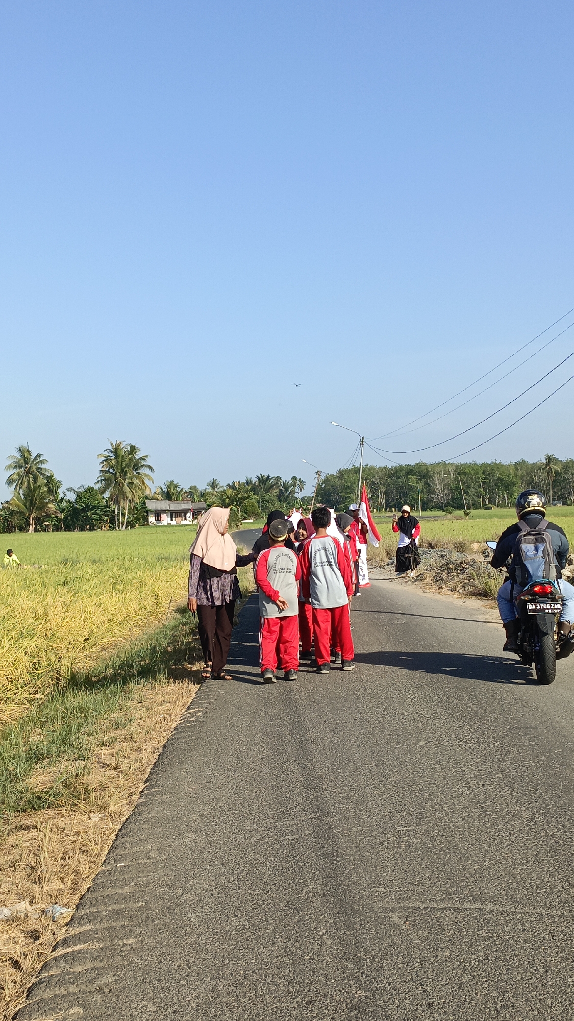 In a scene on a paved road in Batarang, Indonesia, a group of children in red and white uniforms, some carrying Indonesian flags, are walking. A woman wearing a hijab and a patterned tunic is walking alongside them. To the right, a person on a motorcycle with a backpack rides away from the camera. The environment is rural, with a bright blue sky, a field of yellowing crops to the left, and palm trees and other vegetation in the background. A small house with a red roof is visible in the distance. The lighting suggests it is daytime, with the sun casting shadows.