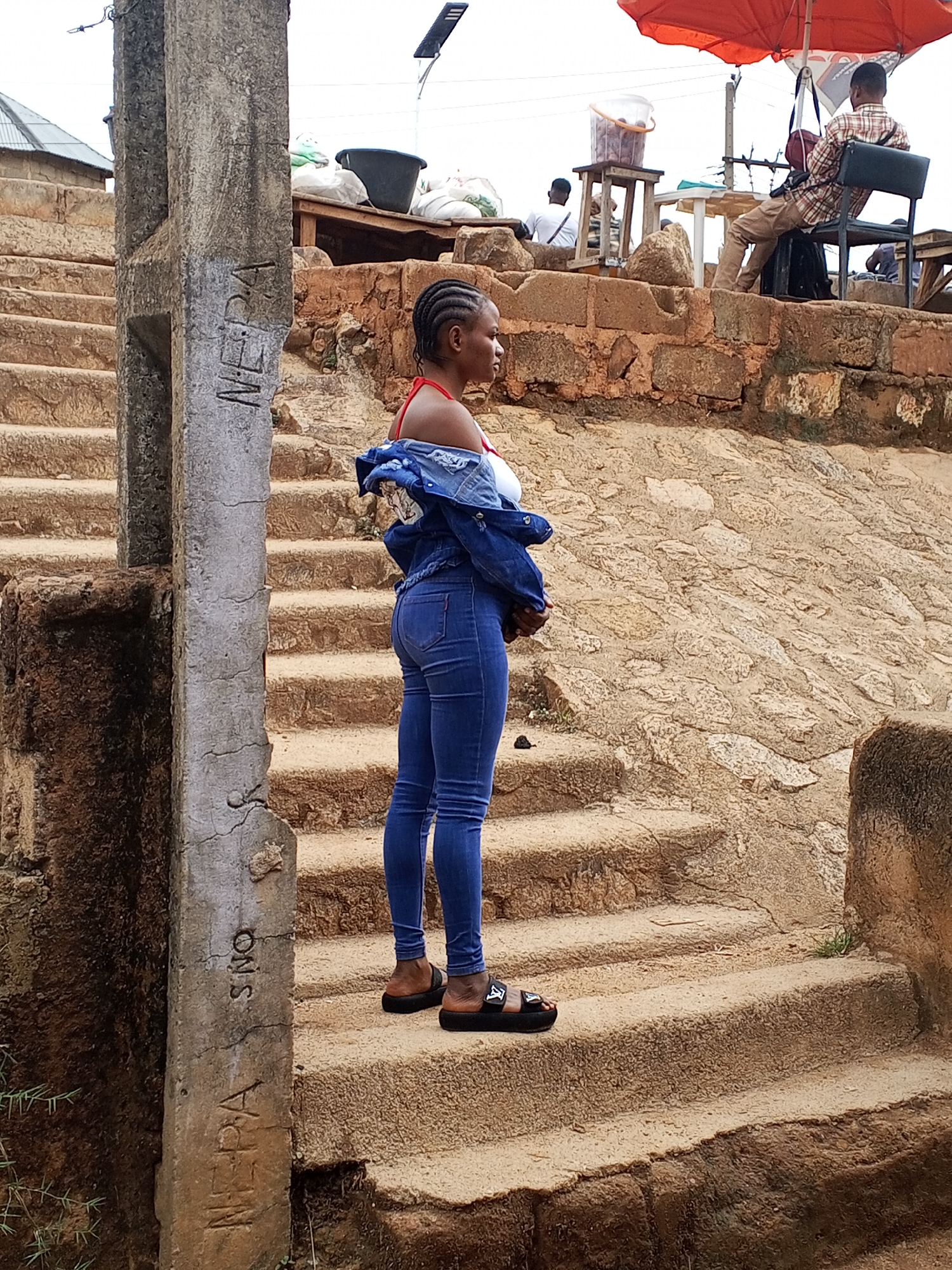 A young woman stands on a set of outdoor stairs in Gwong, Nigeria. She is wearing a white top, blue jeans, a blue denim jacket draped off one shoulder, and black sandals. The stairs appear to be made of stone or concrete and have a worn, weathered look. A weathered concrete pillar with inscribed writing stands to her left. Above and behind her is a raised area with people sitting at tables under an orange umbrella. Other people are present in the background. The environment is outdoors in daylight.
