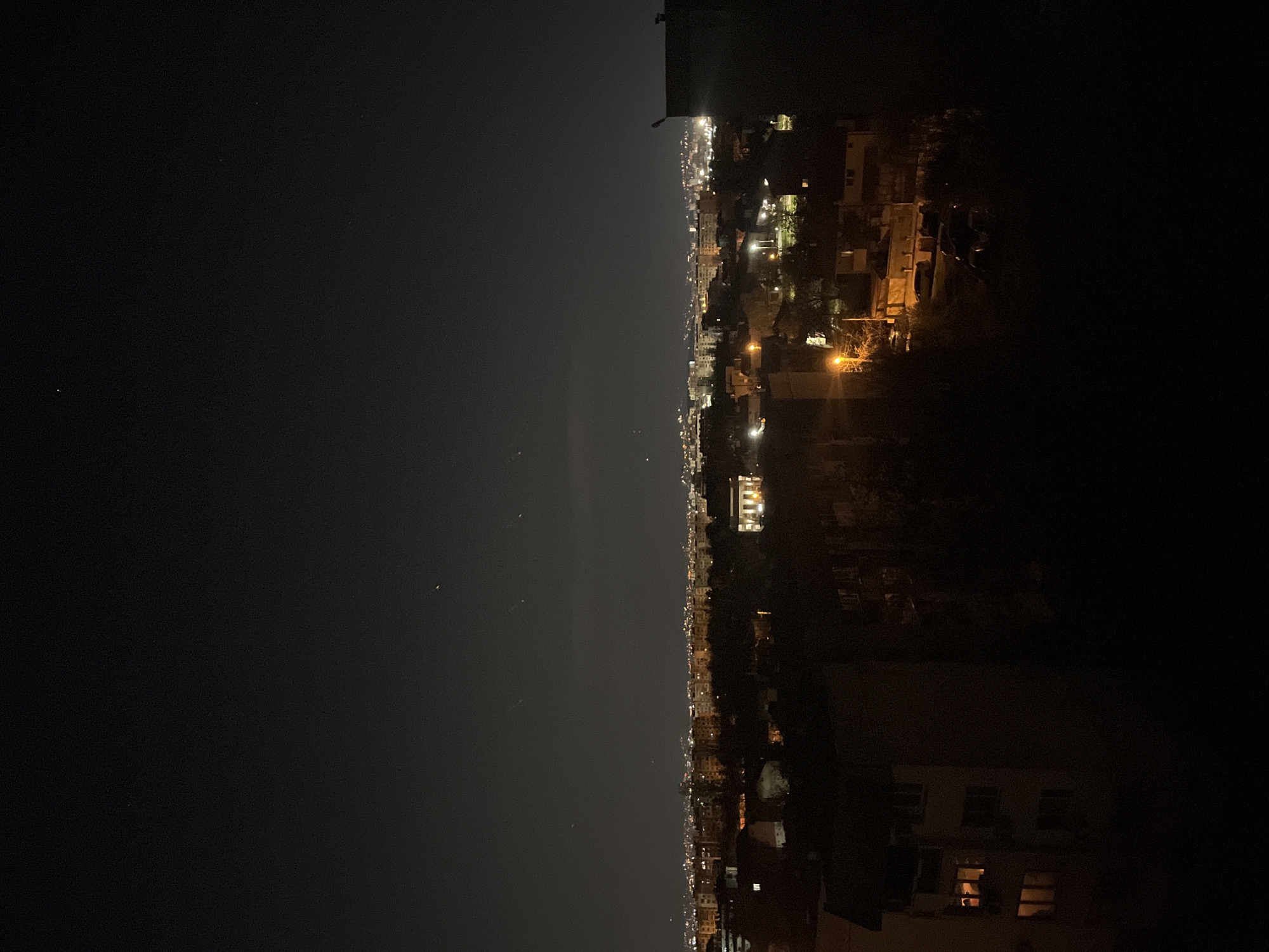 The image depicts a night cityscape in Sofia, Bulgaria. The view is from an elevated position, possibly a high-rise building. In the foreground, structures of nearby buildings are visible, with a few lit windows. Beyond that, a stretch of city buildings extends into the distance, marked by streetlights and illuminated windows. The sky is dark, with a few faintly visible stars.