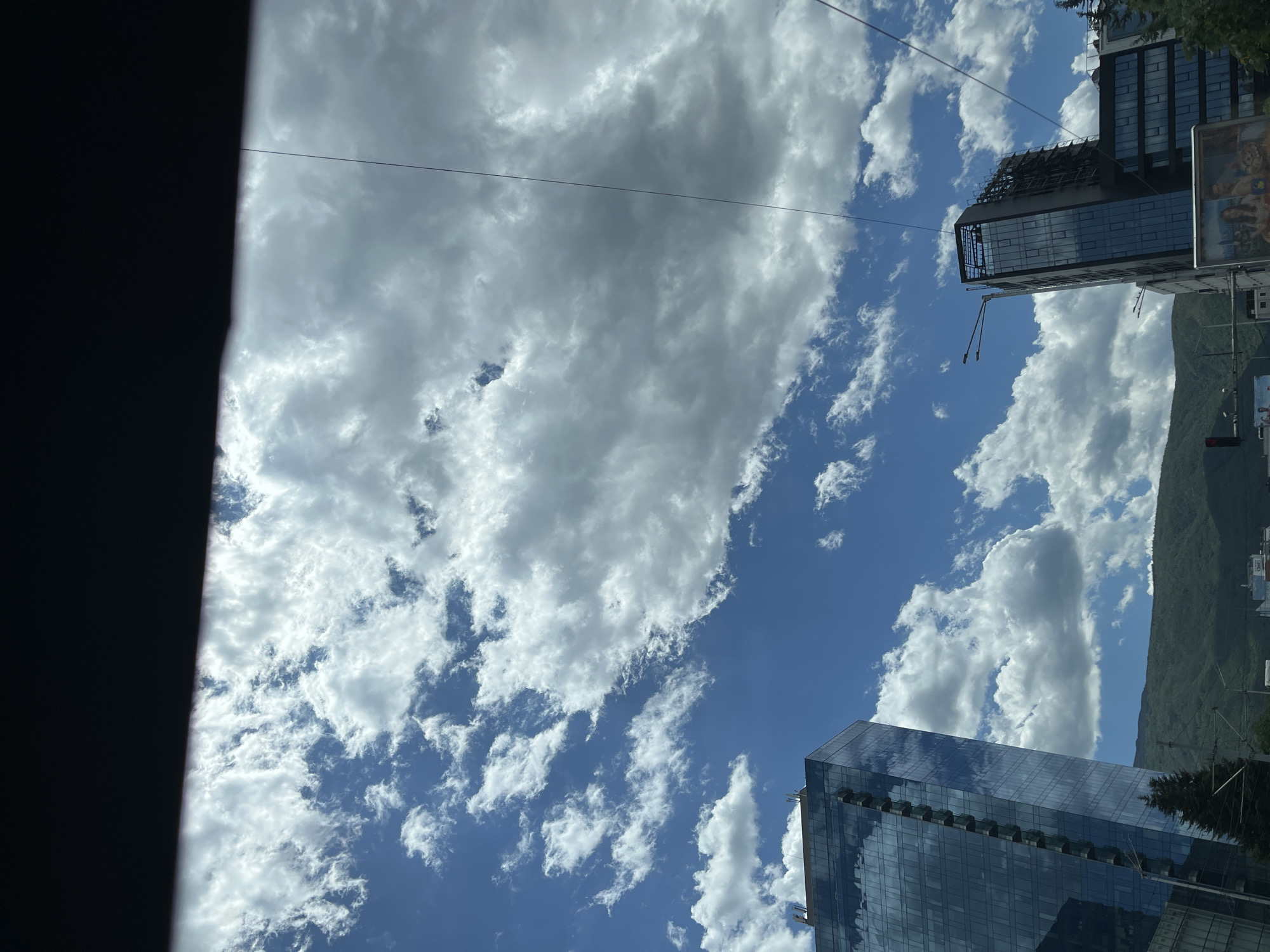 A daytime, low-angle exterior, eye-level shot captures a bright, partly cloudy sky over a city. The top of the frame is dominated by a dark, horizontal element, possibly the interior of a vehicle. The sky is a vibrant blue, with scattered cumulus clouds of varying sizes and densities. On the left, a tall, modern glass-faced skyscraper with reflective surfaces stands prominently. To its right, another contemporary building, also with dark glass facades, is visible, appearing to be under construction or with some unfinished elements at its upper section. In the background, a green, mountainous landscape stretches across the horizon. The lower part of the image shows glimpses of city infrastructure, including traffic lights, streetlights, and advertising billboards. The overall impression is of an urban environment on a clear, dynamic day.