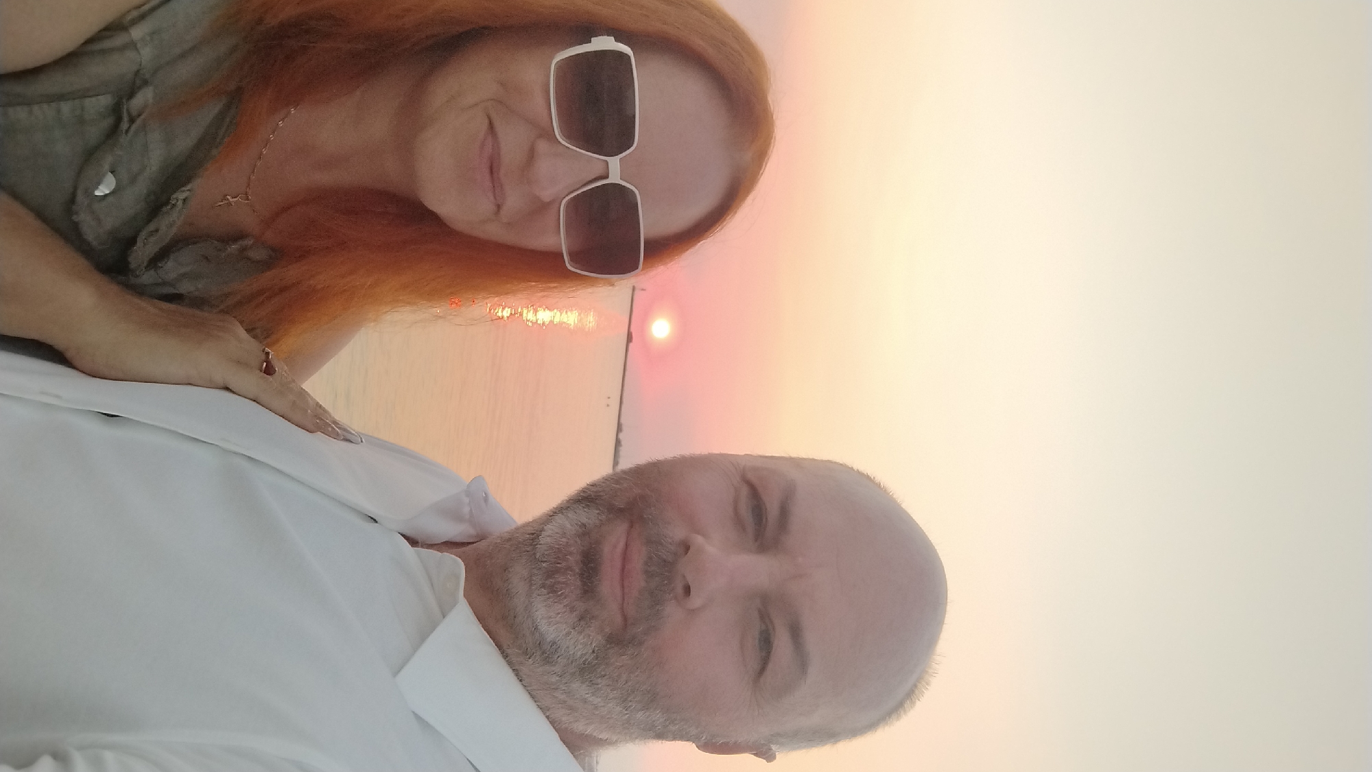 A couple is taking a selfie at sunset near the beach in Neoi Epivates, Greece. The woman on the left has long, red hair and is wearing large, white-rimmed sunglasses and a green vest. She is also wearing a delicate necklace with a cross pendant. The man on the right has short, grey hair, a grey beard, and is wearing a white collared shirt. His left hand is placed on the woman's arm. The sun is low on the horizon, casting a warm, orange glow over the water, which reflects the colors of the sunset. The sky is a pale, creamy color.