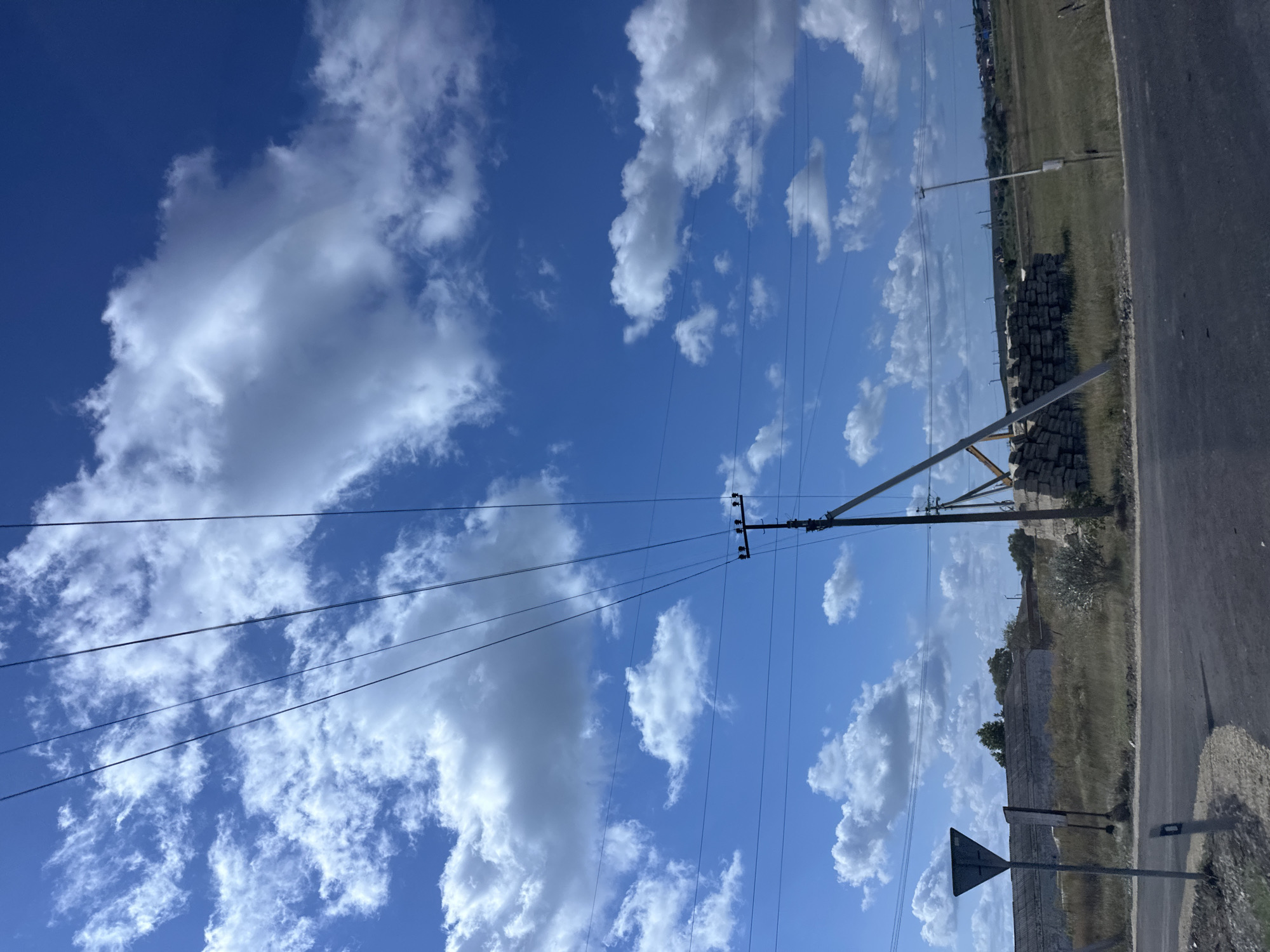 The image depicts a rural or semi-rural roadside scene in Peresyp, Russia, under a bright blue sky with scattered white clouds. Several power lines stretch diagonally across the frame, converging towards a single wooden utility pole with insulators. In the foreground, a paved road curves to the right, with a gravel shoulder. A triangular road sign is visible on the left side of the road, and a short gray post with a white marking is next to it. To the right of the utility pole, a stack of construction materials, possibly bricks or stones, is piled up. Beyond the road and construction materials, the landscape is covered in dry grass and scrub brush. In the middle ground, a long, low building with a corrugated roof is visible, and further in the distance, a few scattered houses and trees can be seen. The overall impression is one of an undeveloped or developing area. There are no people or animals present in the image.