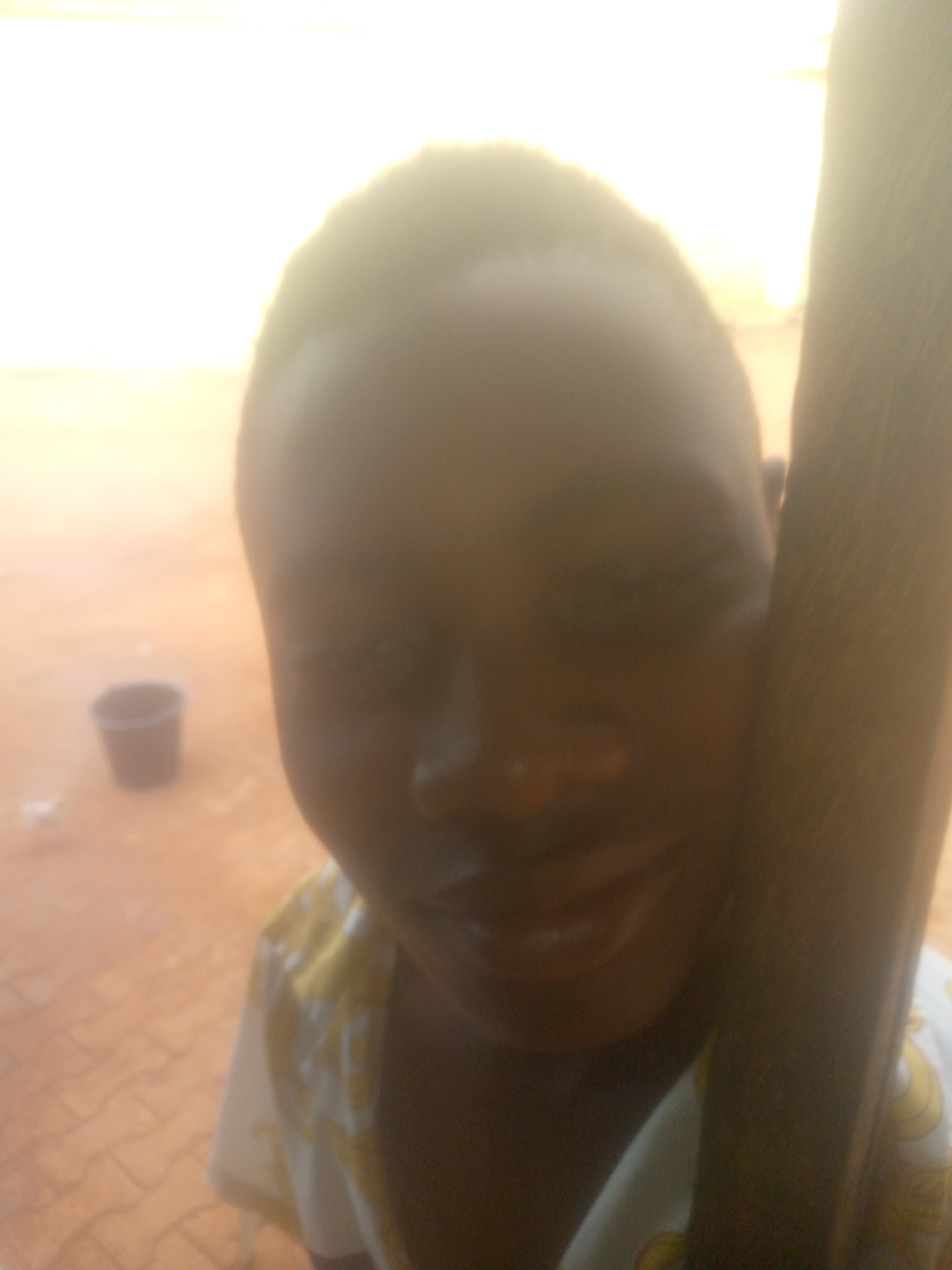 The image shows a young person with dark skin, likely a child or youth, looking directly at the camera with a subtle smile. Their head and upper body fill a significant portion of the frame, with the light source coming from behind and to the left, causing their features to be somewhat shadowed. They are wearing a light-colored shirt, possibly white with yellow or gold patterns visible on the shoulders.
To the right, a dark, vertical object, appearing to be a wooden pole or post, runs along the edge of the frame. The background is extremely bright and overexposed, indicating strong sunlight, making it difficult to discern specific details. The ground is a reddish-brown color, which could be dirt or pavement, and a dark, cylindrical bucket is visible in the distant left background. The strong backlighting creates a bright halo effect around the person's head. The scene is outdoors during bright daytime. No text is visible.