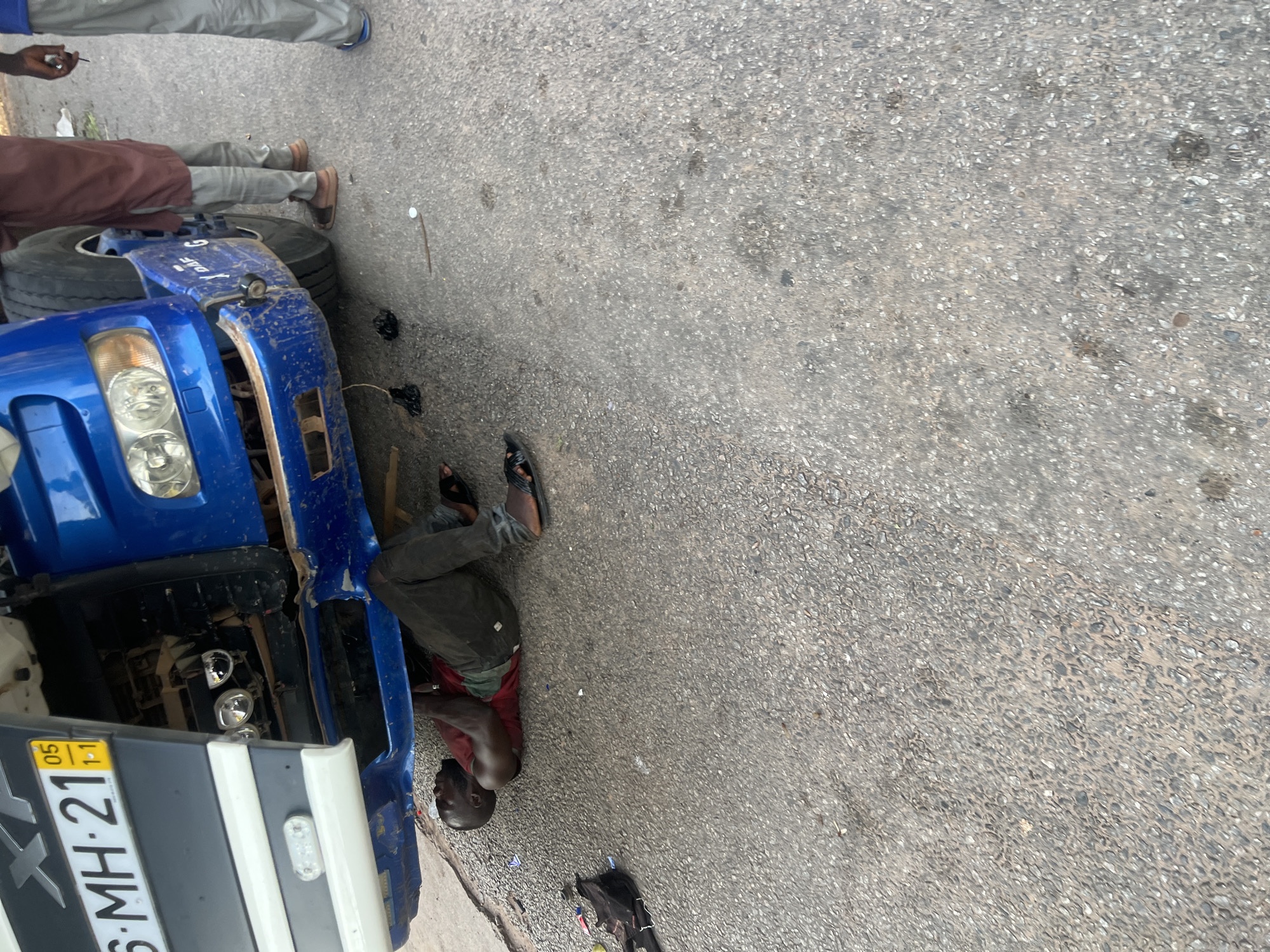 The image captures a roadside repair scene in Jalingo, Nigeria. A blue heavy-duty truck, identified by its visible "DAF G" logo and a partially visible "XF" on its grill, has its cab tilted forward, exposing the engine compartment for maintenance. The truck bears a Nigerian license plate "6-MH-21," with a small yellow sticker indicating "05 1-1," possibly signifying an expiration or registration detail. The front right side of the truck's bumper is visibly damaged and splattered with mud.
An adult male, with dark skin and a bald head, is lying on his back directly on the asphalt road surface beneath the front of the truck. He is wearing a reddish-brown t-shirt, dark trousers, and dark flip-flops, with his arms raised and hands engaged with components under the vehicle, actively performing a repair.
To the right of the truck, two other adult males are partially visible, standing on the asphalt. One individual, closer to the truck, is wearing a reddish-brown long-sleeved garment and light grey trousers, with brown sandals. The second individual, further to the right, is largely obscured, with only his lower body visible, wearing light grey trousers and blue flip-flops, and holding a small dark object in his right hand. Both individuals appear to be observing the ongoing repair.
The road surface is asphalt, a light greyish color with visible dark aggregate, showing wear and minor scattered debris including small dark plastic items, a white cap-like object, and a stick-like object on the ground near the truck's front wheel. The scene is outdoors, under daylight conditions, focusing entirely on the vehicle and the individuals present.