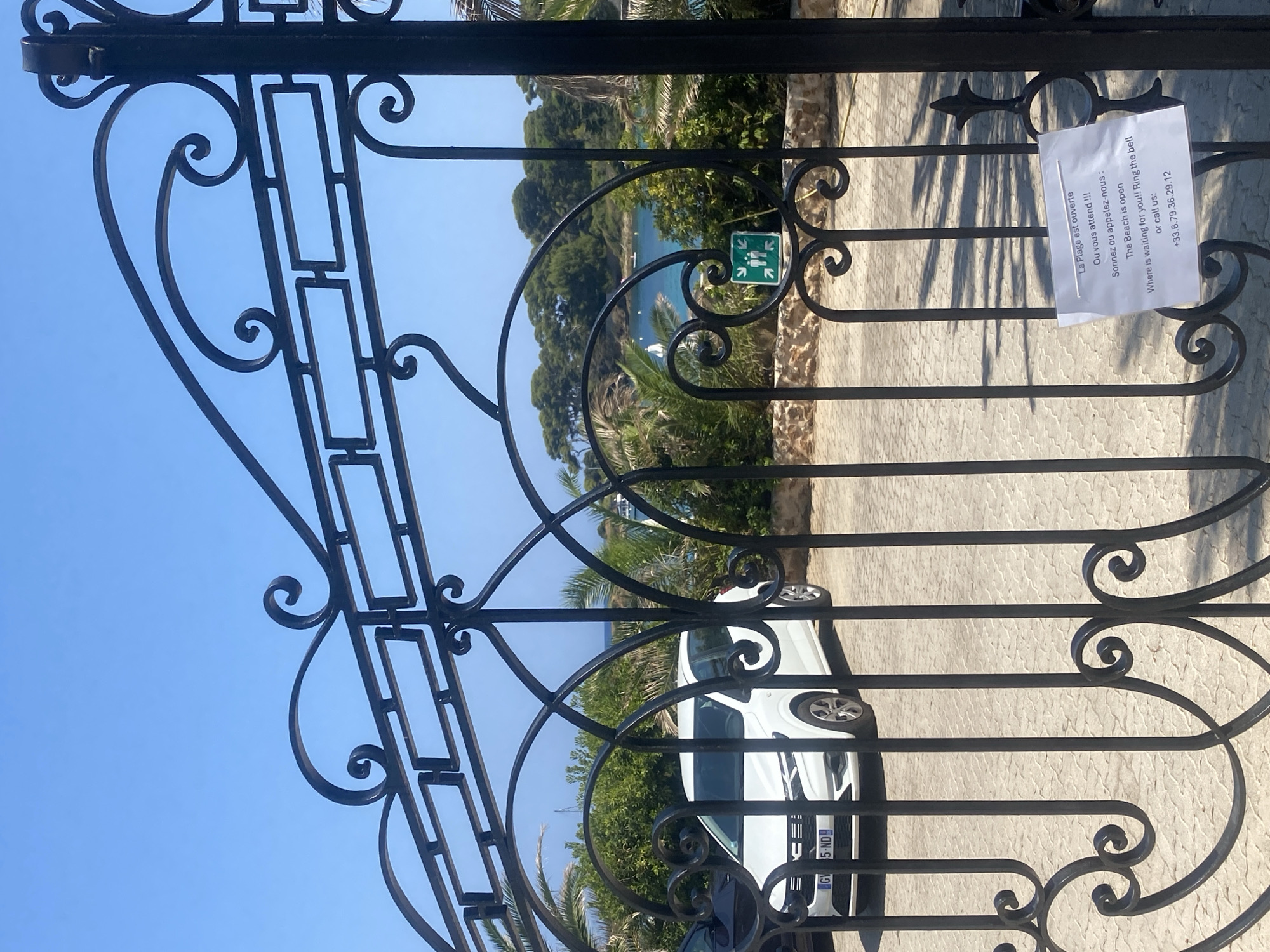 This is a vertical, eye-level outdoor shot of a black ornate metal gate with scrolled accents. Through the gate, a white car is parked on a paved area. Beyond the car, lush green foliage and palm trees line the coast of a bright blue body of water, with landmasses in the distance. The sky is clear and blue. A white sign is attached to the gate with text in French and English. The French text reads "La Plage est ouverte. Ou vous attend !!!". The English text reads "The Beach is open. Where is waiting for you!! Ring the bell or call us: +33.6.79.36.29.12". The location appears to be a beach resort or private property in Antibes, France, given the context of the French language and the Mediterranean scenery.