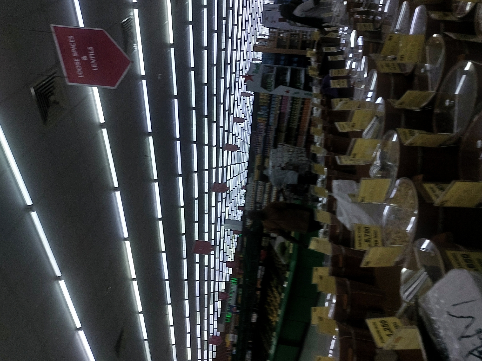 This image depicts the interior of a supermarket or grocery store in Dar es-Salaam, Tanzania, characterized by a bright, uniform overhead lighting system. The ceiling is fitted with numerous parallel rows of long, white fluorescent light fixtures and several square air vents.
A red, downward-pointing hexagonal sign hangs from the ceiling in the upper right quadrant, clearly labeled "LOOSE SPICES & LENTILS" in white capital letters. Other smaller, rectangular hanging signs are visible further back within the store.
The foreground and mid-ground are dominated by an array of large, open wooden or plastic bins containing various bulk food items. Many yellow rectangular price and information tags are displayed in front of these bins. Notable tags include one reading "PUMPKIN SEEDS 1,250" and another stating "UNSALTED NUTS 5,700 PER 100G." Several silver scoops are placed within the bins, indicating self-service. A white plastic bag is visible near some bins.
In the mid-ground, extending from the left, is a long, green display unit, likely for fresh produce, on which indistinct dark items are visible.
Several blurry and indistinct human figures are present within the store. One person, wearing a dark brown or tan long-sleeved garment, is partially visible from the back, standing near the green display unit and facing right. Further back, another individual appears to be wearing a light-colored top. In the far background right, a person with dark hair and a light-colored top is also visible.
The background features tall shelving units stocked with numerous packaged goods, appearing as colorful rows. A white and green promotional display, possibly featuring a star logo, stands near these shelves on the right. The overall environment appears well-maintained and commercially organized.