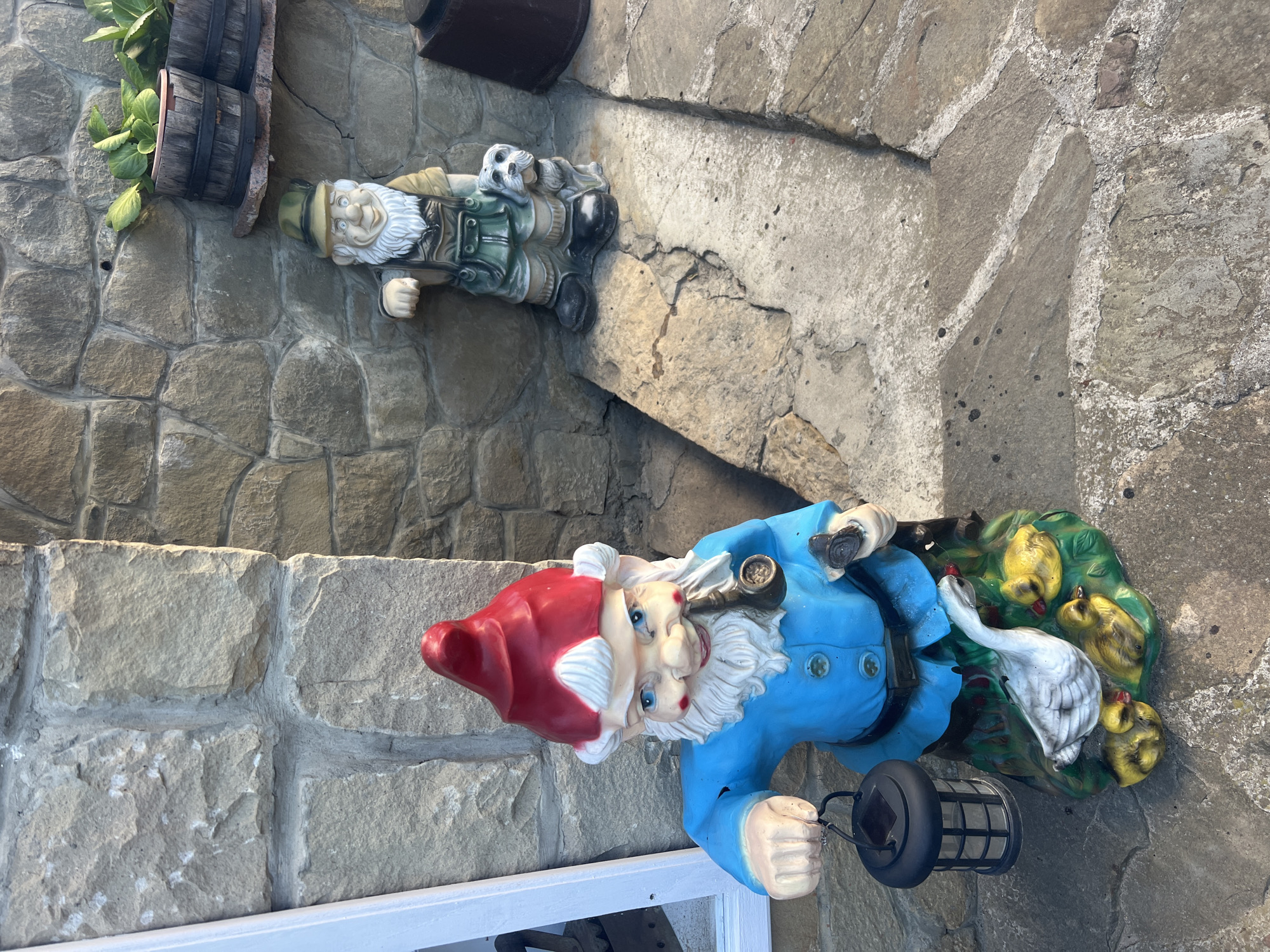 Two garden gnome statues are positioned outdoors. The gnome in the foreground is larger and more brightly colored, wearing a blue jacket, a red pointed hat, and a white beard. It holds a black lantern in its left hand and a smoking pipe in its right. The gnome is standing on a base featuring a white goose and three yellow ducklings. Behind this gnome, a second, smaller gnome statue is visible. This gnome wears a green hat, a green and tan outfit, and is accompanied by a small white dog statue. Both gnomes are placed on a stone pathway and a stone wall forms the backdrop. The overall scene suggests a decorative outdoor setting. The location is identified as Yaremche, Ukraine.