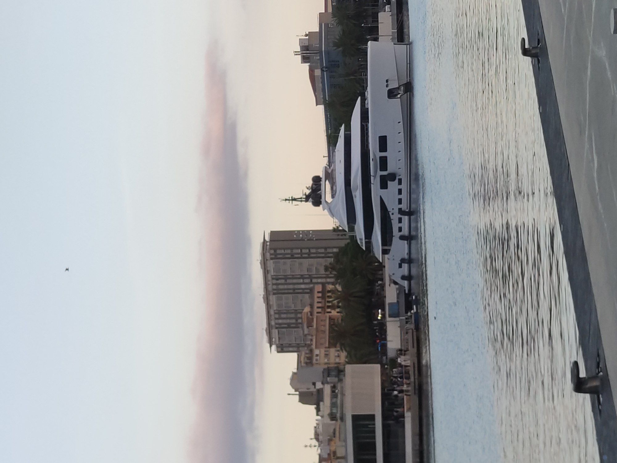 A large, white luxury yacht is docked in a harbor in Barcelona, Spain, during dusk. The yacht is the dominant subject, positioned prominently in the mid-ground and right of the frame. It features multiple decks, sleek lines, and numerous windows.
In the background, the Barcelona skyline is visible, with a mix of modern and older buildings. A tall, rectangular building with many windows stands out, likely a hotel or apartment complex. Adjacent to it are other buildings of varying heights and architectural styles, some appearing older with more traditional facades. Palm trees are scattered among the buildings, indicating a Mediterranean climate.
The water in the foreground is calm, reflecting the soft light of the sky. There is a dark, textured surface in the immediate foreground, possibly a dock or pier, with a metal mooring bollard.
A few people can be seen on a lighter-colored platform or walkway attached to the shore near some of the background buildings, suggesting a public area or promenade. The sky is a gradient of pale blue and light pink, with some wispy clouds, characteristic of sunset or sunrise. A solitary bird is visible in the upper sky. The overall scene conveys a peaceful, urban waterfront atmosphere.