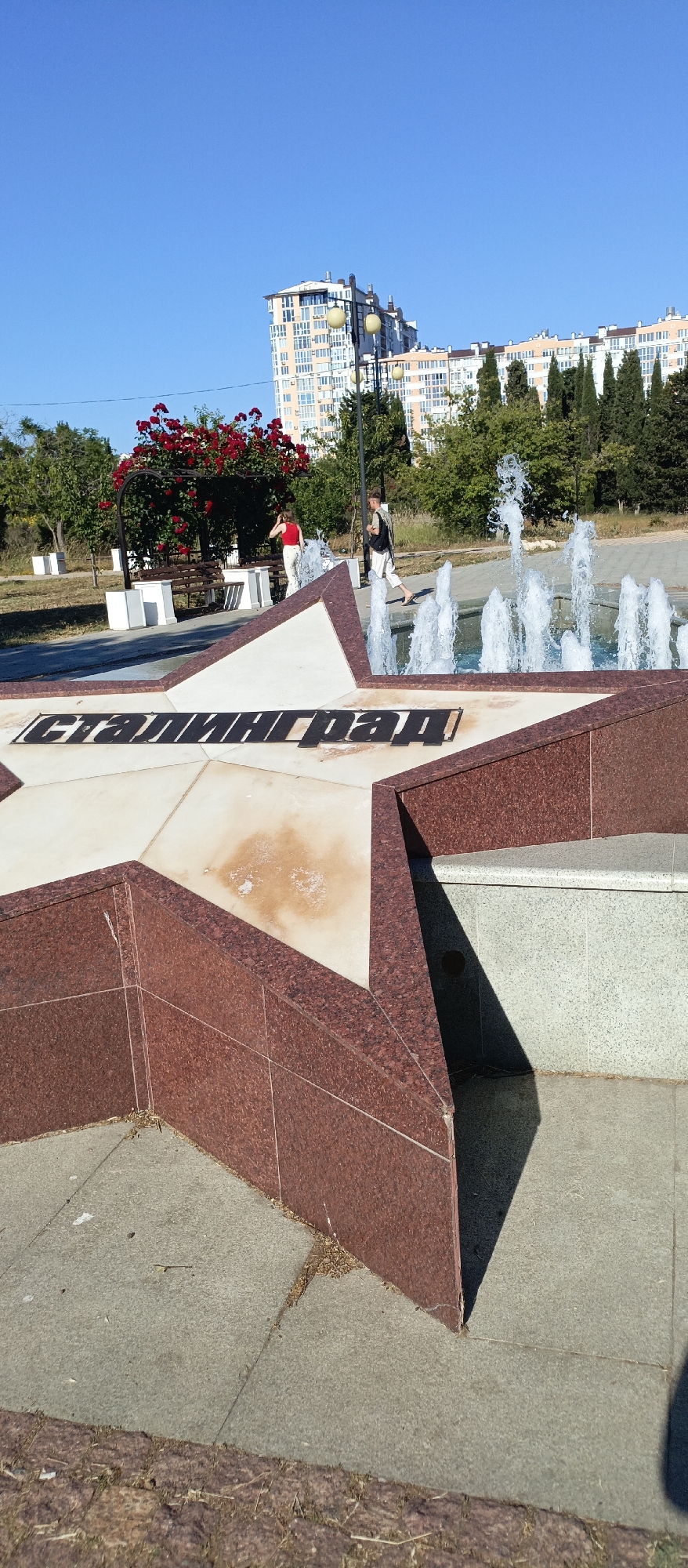 In the city of Unknown, Unknown, on a sunny day, a monument in the shape of a star is the central focus. The star monument features the inscription "Сталинград" in black lettering across its surface. In the background, a fountain sprays water upwards, and two individuals are present near the fountain. One person, wearing a red top and light-colored pants, stands near a park bench under a vibrant red rose bush. The other individual, wearing a dark backpack and light-colored pants, walks towards the fountain. Behind them, multi-story buildings with light-colored facades and balconies dominate the skyline, interspersed with trees. The ground is paved with concrete slabs, and dry grass and leaves are scattered around the monument. The sky is a clear, bright blue.
