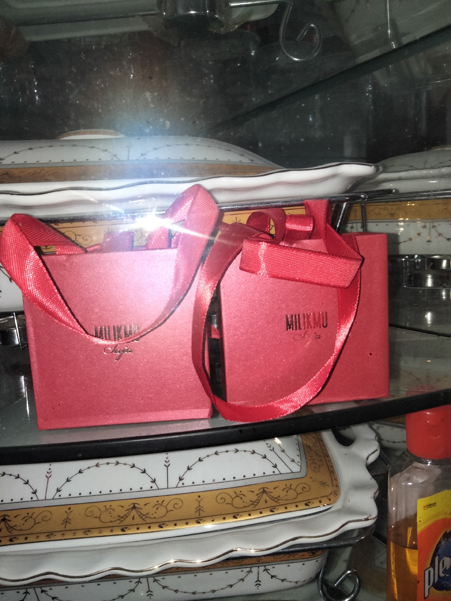 The image shows two small red gift boxes with red ribbon handles, likely sitting on a shelf in a display cabinet. The boxes have gold lettering that appears to read "MILIKMU" and "Suci". Below the boxes, there are decorative white and gold ceramic platters. To the right of the platters, a small bottle of what appears to be a liquid with an orange cap is visible. The scene is lit by artificial light, possibly from within the cabinet, creating some reflections on the glass surfaces. The overall impression is of a display of small gifts or decorative items, perhaps in a home or a shop setting in Payagaboh, Indonesia. There is no visible indication of the time of day or weather conditions.