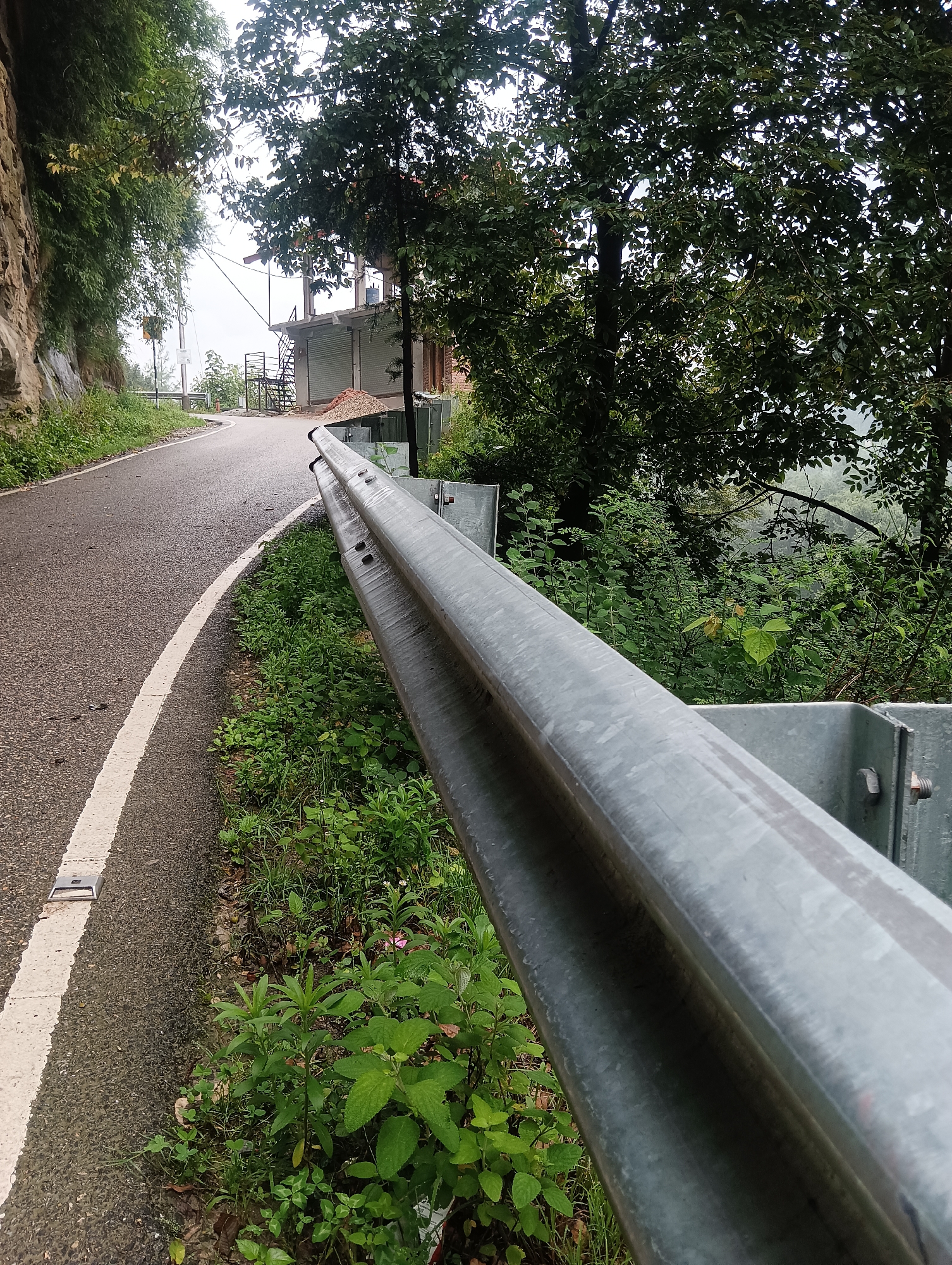 The image depicts a roadside scene on a paved road in the vicinity of Goshal, India. The road curves to the left, bordered by a metal guardrail on the right and a rocky embankment with vegetation on the left. The guardrail is constructed of corrugated metal and is supported by metal posts. Lush green vegetation, including various plants and trees, grows along the side of the road and on the embankment. In the background, there is a partially constructed building with scaffolding and what appear to be rolled shutters. The sky is overcast, suggesting a cloudy or misty day. The lighting is diffused, with no strong shadows. There are no people or animals visible in the image. The overall scene is one of a rural or mountainous road under construction or development.