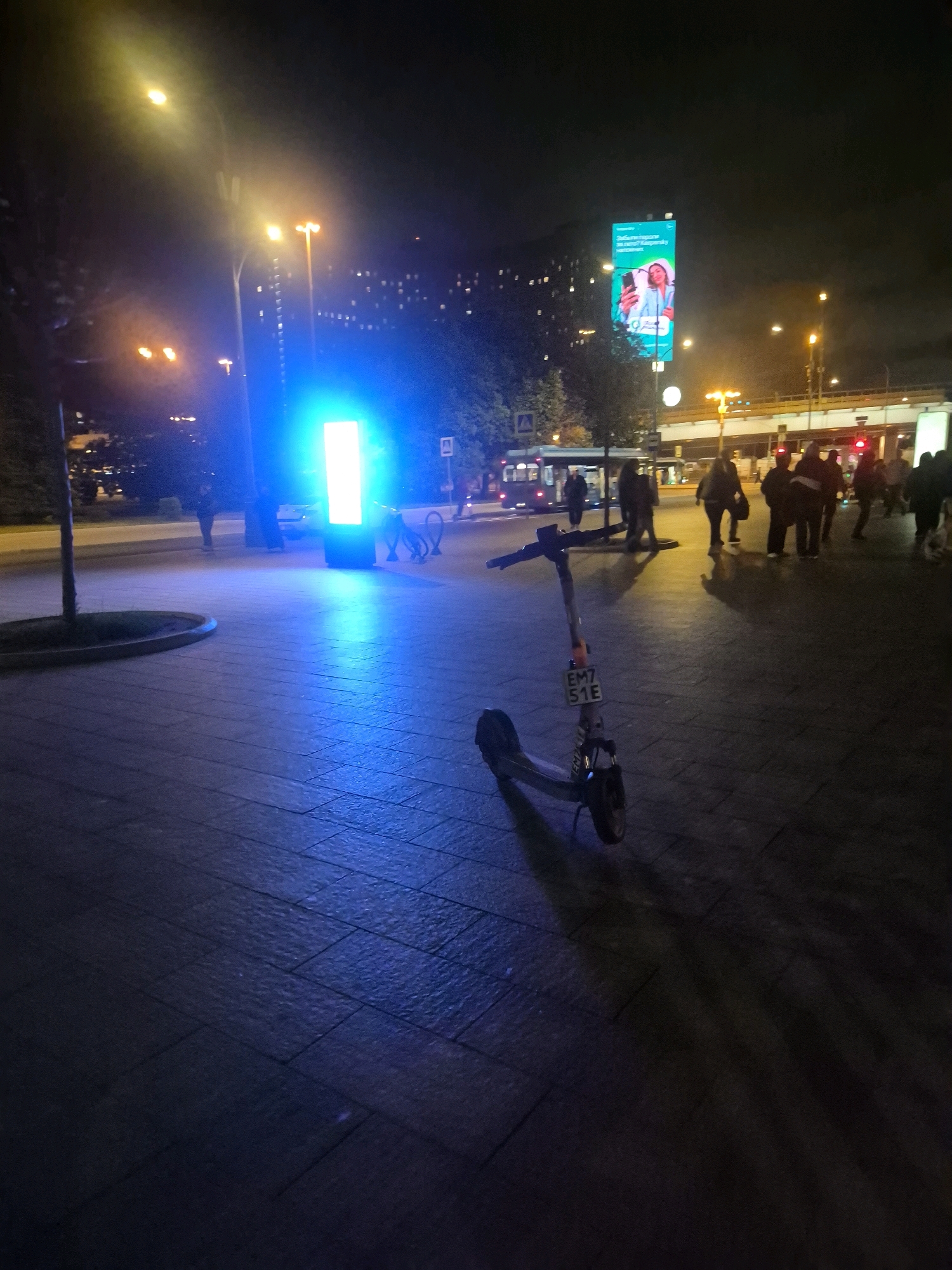 The image depicts a nighttime scene on a pedestrian walkway in Moscow, Russia. A gray electric scooter is prominently positioned in the foreground, angled towards the right. The scooter has a license plate reading "EM7 51E." In the middle ground, a bright blue illuminated pillar or structure casts a strong blue light on the pavement. Several pedestrians are visible in the background, walking and standing. To the left, a tree with sparse foliage stands. Further in the background, city lights and buildings are discernible, along with a large billboard displaying an advertisement. Streetlights with yellow and white illumination are also present. The ground is paved with stone tiles.