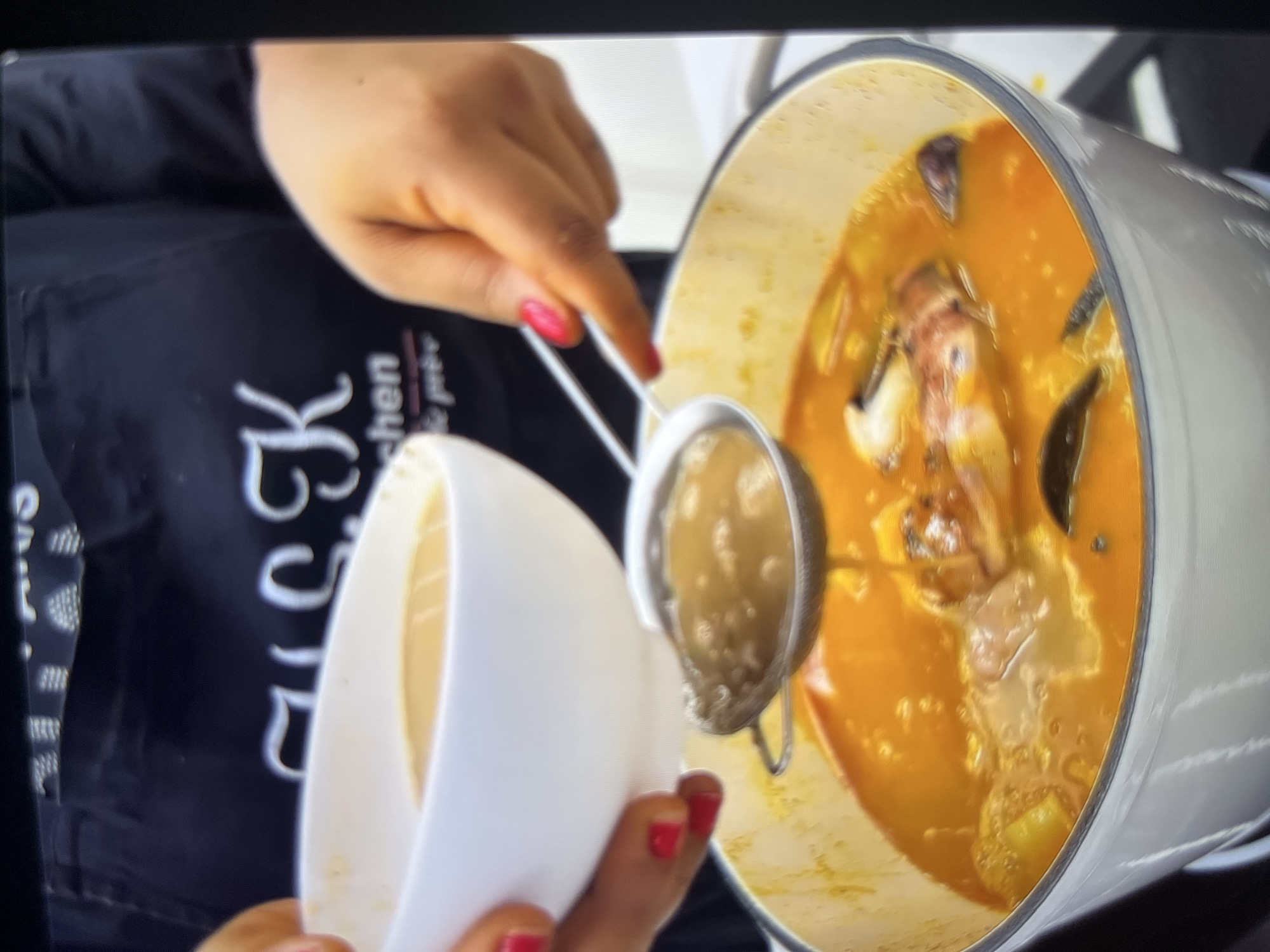 In the foreground, a person with bright pink nail polish is pouring a liquid from a white bowl through a small sieve into a white pot of soup. The soup is a vibrant orange color and appears to contain pieces of seafood, possibly shrimp, and some vegetables. The pot is on a stovetop. The person is wearing a black apron with white lettering that appears to say "Kitchen". The background is blurred, but the overall impression is of a cooking scene. The provided crops do not offer any discernible details that would allow for a specific location in Abuja, Nigeria.