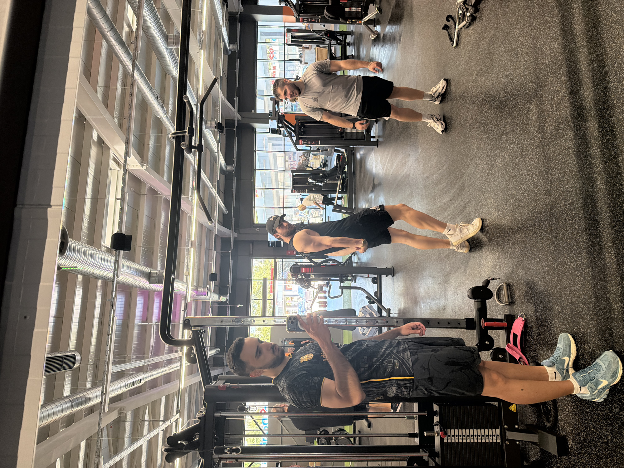 Three men are in a gym with various fitness machines. The man closest to the camera is standing near a cable machine, holding a smartphone. The second man is leaning over a weight bench. The third man is standing upright and appears to be smiling. The gym has a high ceiling with exposed ventilation ducts. The location is not identifiable as Plovdiv, Bulgaria without further information.