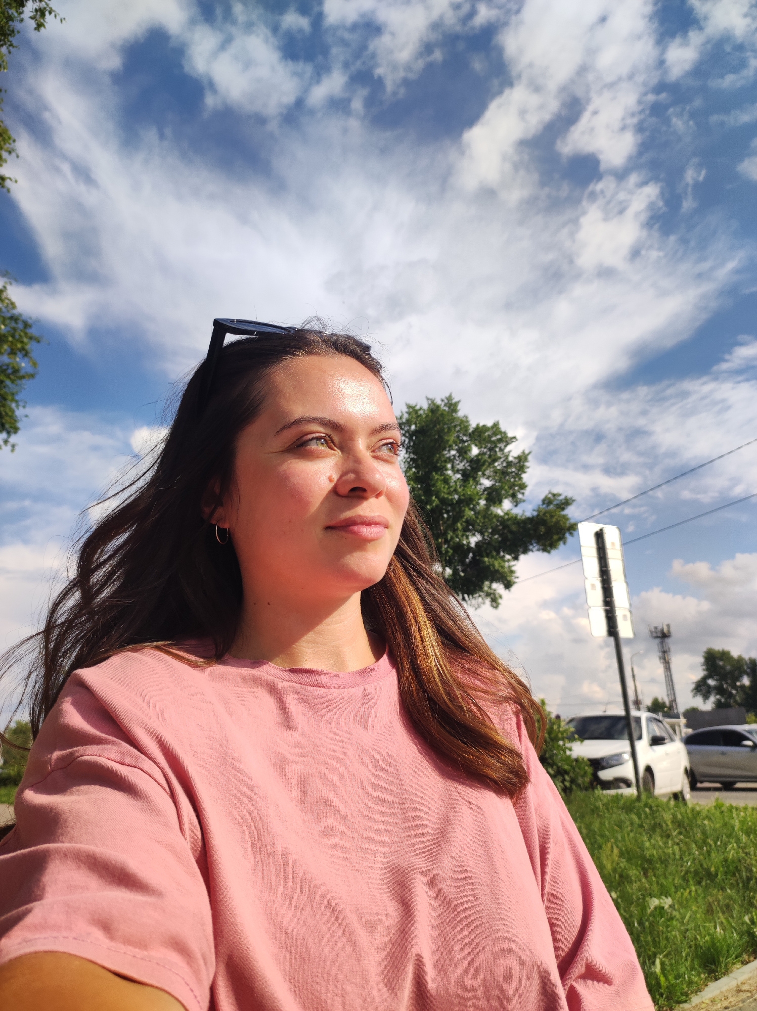 A young woman with long, dark brown hair is looking to her right, her face illuminated by bright sunlight. She is wearing a pink t-shirt and has a pair of dark sunglasses resting on top of her head. Her expression is calm and contemplative. The background features a bright blue sky with scattered white clouds, indicating fair weather. To the right of the woman, there is a street with cars, a traffic sign, and some greenery, suggesting an outdoor urban or suburban setting. The lighting indicates it is daytime, likely late afternoon due to the angle of the sun. No visible text is present in the image. The context of Chaikovsky, Russia, places the scene within that specific geographical location.