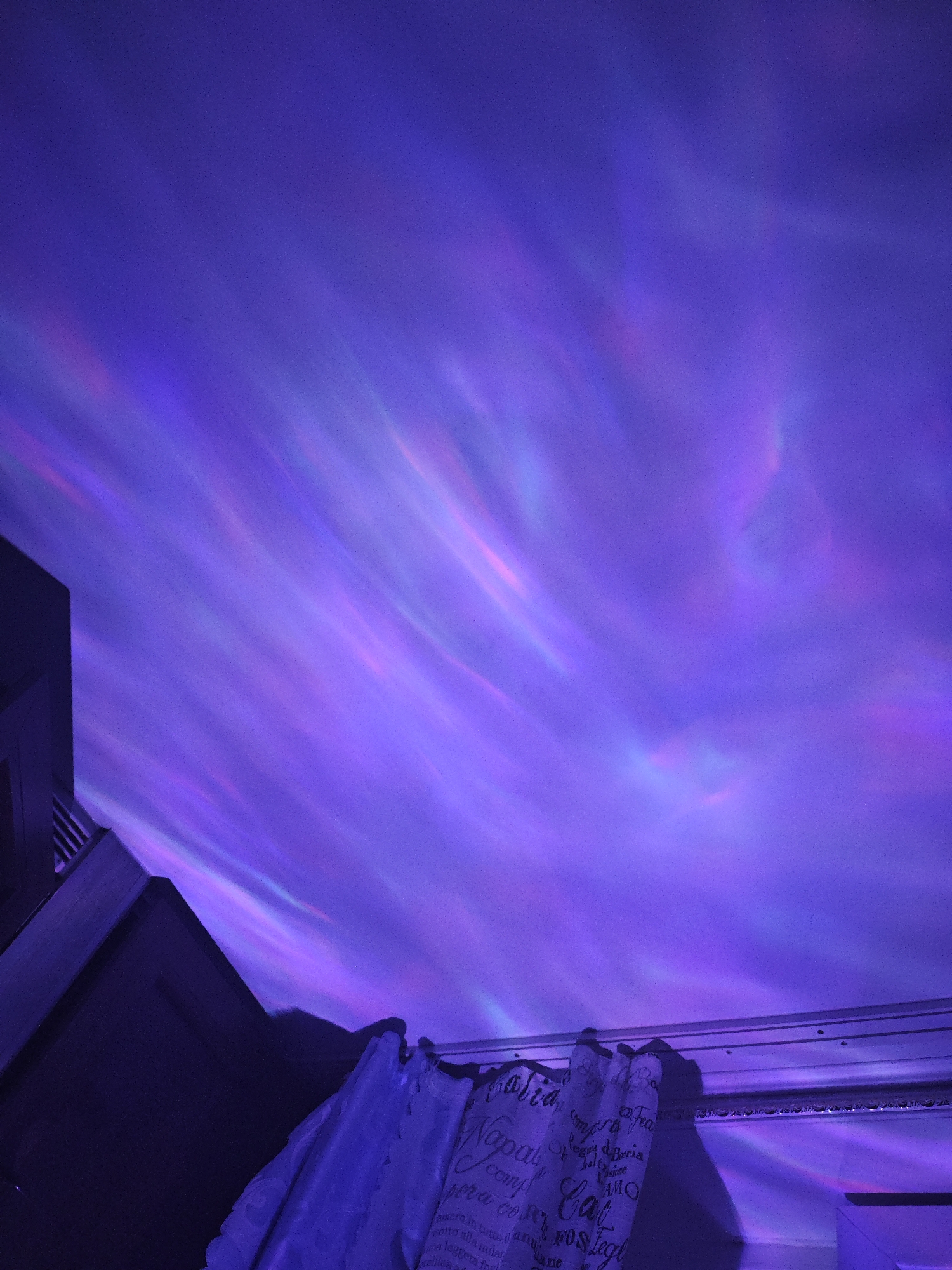 The image shows a ceiling illuminated by purple and pink lights that create a wavy, aurora-like effect. In the lower left and right corners, dark architectural elements are visible, possibly part of a room's structure. A draped fabric with text is present in the lower center and right. The overall impression is an indoor space with atmospheric lighting. There are no people or animals visible in the image.