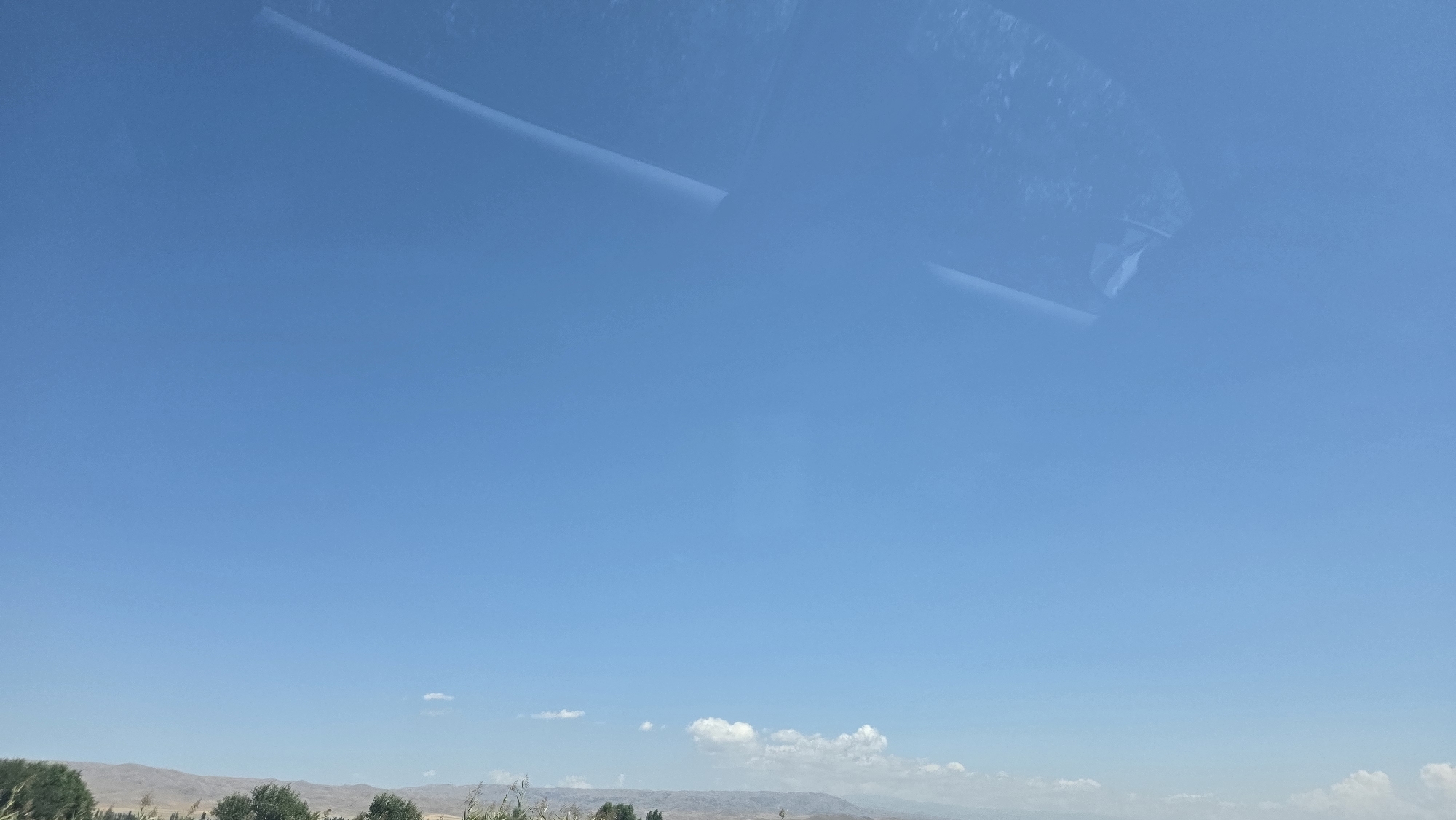 The image shows a clear blue sky with a few scattered white clouds on the horizon. The lower portion of the image depicts a landscape with dry, rolling hills. In the foreground, there are some trees and vegetation. There are no people or significant human activity visible. The scene appears to be an outdoor environment, possibly in a rural or semi-arid area. The upper part of the image has some reflections on what could be a window, suggesting the photo may have been taken from inside a vehicle. The location is identified as Krasnaya Rechka, Kyrgyzstan.