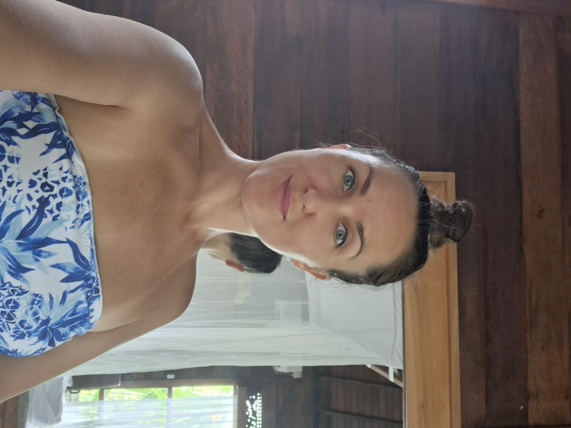 A woman is depicted in a close-up, half-length portrait, likely a selfie, taken indoors within a rustic room. She has dark hair styled in a bun, light blue eyes, and fair skin with a slight redness on her cheeks and forehead. Her lips are softly closed, forming a gentle, subtle smile, and she looks directly at the camera. She is wearing a strapless garment, possibly a wrap dress or towel, patterned with white and blue tropical or botanical motifs, including what appear to be pineapples.
The setting features warm, wooden plank walls. Behind her, a reflection of the back of her head is visible in what appears to be a large mirror or reflective surface. Below this reflection, a window frames a vibrant outdoor scene of lush green foliage, indicating a tropical environment. A white, sheer mosquito net is draped in the background, suggesting a bed canopy. Part of a wooden structure, possibly a bed frame, is also visible. The natural, bright lighting from the window suggests it is daytime. The overall impression is one of relaxation in a tropical, possibly traditional, accommodation, consistent with the specified location of Ubud District, Indonesia. No text is visible in the image.