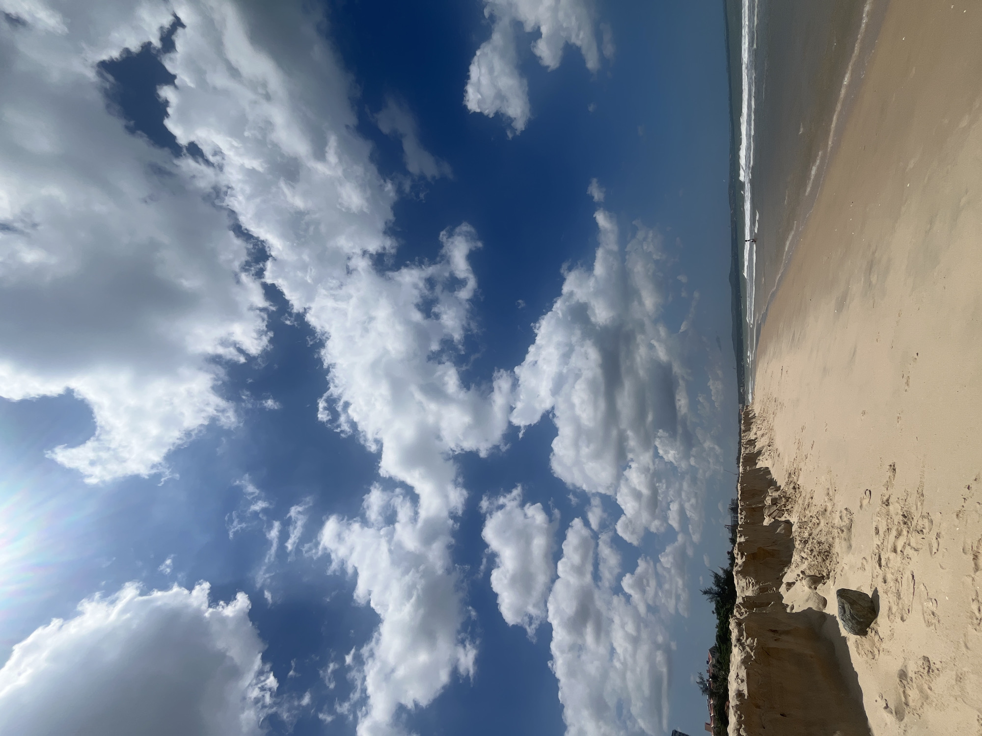 This is a landscape photograph of a sandy beach with a view of the ocean under a partly cloudy sky. The image is oriented vertically, giving a sense of looking up towards the sky and then down towards the beach.
The foreground of the image is dominated by a sandy beach, which appears to be eroded in some areas, revealing layered sand. A few footprints are visible in the sand, suggesting recent activity. A dark rock is also present in the sand.
Further back, the ocean is visible with gentle waves rolling onto the shore. A thin line of white foam marks the edge of the water. In the distance, along the horizon, there are some indistinct buildings and trees, possibly indicating a coastal town or resort. A lone figure can be seen walking near the water's edge.
The sky is a deep blue, dotted with numerous white, fluffy clouds. Some of these clouds are quite large and detailed, while others are more wispy. The sun appears to be out, as indicated by the bright areas and lens flare in the upper left portion of the image. The lighting suggests it is daytime, likely mid-morning or afternoon.
The overall mood of the image is peaceful and serene, evoking a sense of a calm day at the beach. There is no visible text in the image. The location cues, such as the type of sand, vegetation, and distant architecture, along with the provided location context, suggest this is Phu Thuy Ward, Vietnam.