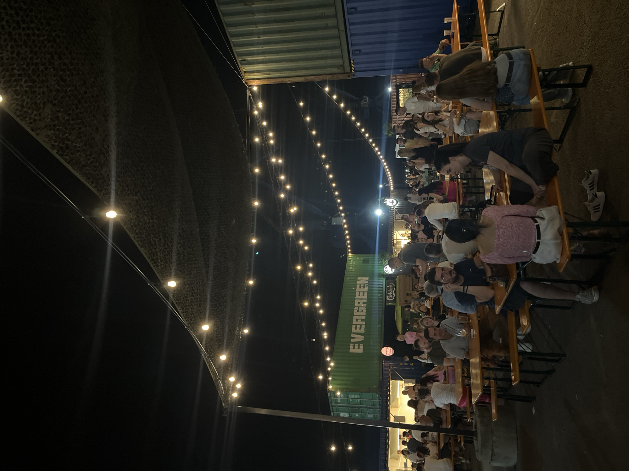 The image depicts an outdoor seating area at night, illuminated by string lights. A large group of people are seated at long, communal tables, some eating and drinking, while others are engaged in conversation. In the background, shipping containers have been repurposed for what appears to be a food or drink establishment. One container is painted green and has the word "EVERGREEN" in white lettering. Another container, to the right, is blue. The overall atmosphere suggests a lively, casual gathering place. The scene is located at Beton Hala in Belgrade, Serbia.