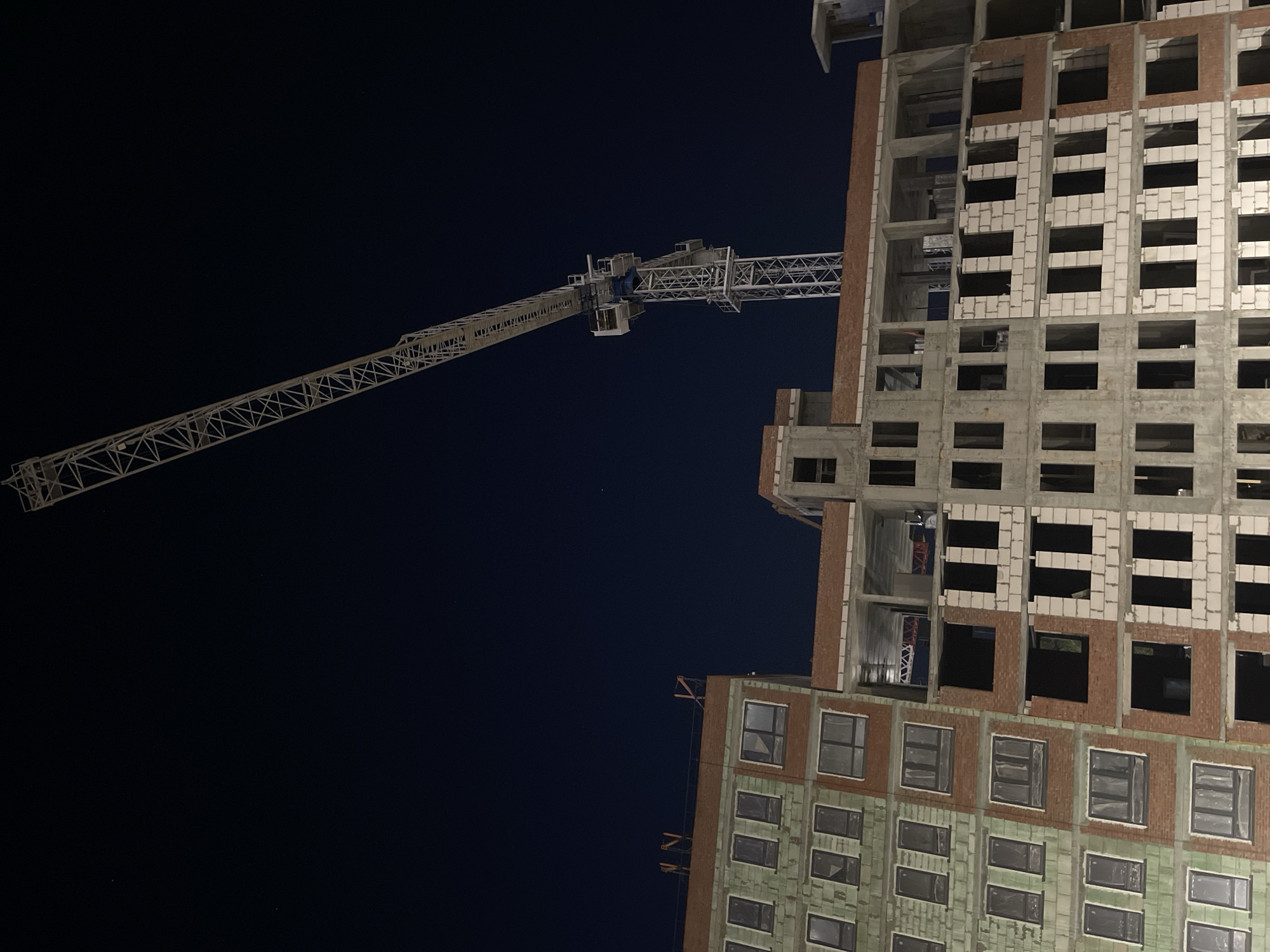 The image depicts a construction site at night, dominated by a towering crane and a multi-story building under construction. The upper and left areas of the frame are filled with a deep, dark night sky, indicating a clear, cloudless evening.
A large metal tower crane extends diagonally across the middle of the frame. Its long, latticed jib stretches from the bottom-left towards the upper-right, with its central mast and operator's cabin visible in the mid-right. The crane is subtly illuminated, highlighting its metallic structure against the dark background.
To the right, a multi-level building is in various stages of construction. The upper sections display exposed concrete frameworks, interspersed with light-colored aerated concrete blocks and reddish-brown brickwork forming the walls. Many window openings are dark and empty. Lower down, towards the bottom-right, sections of the building appear more complete, featuring a greenish-gray facade and installed, dark-paned windows. The juxtaposition of raw construction materials with more finished sections suggests active development.
The scene conveys an industrial atmosphere, capturing a moment of stillness at a busy construction project during non-working hours. No people or identifiable text are visible in the image.