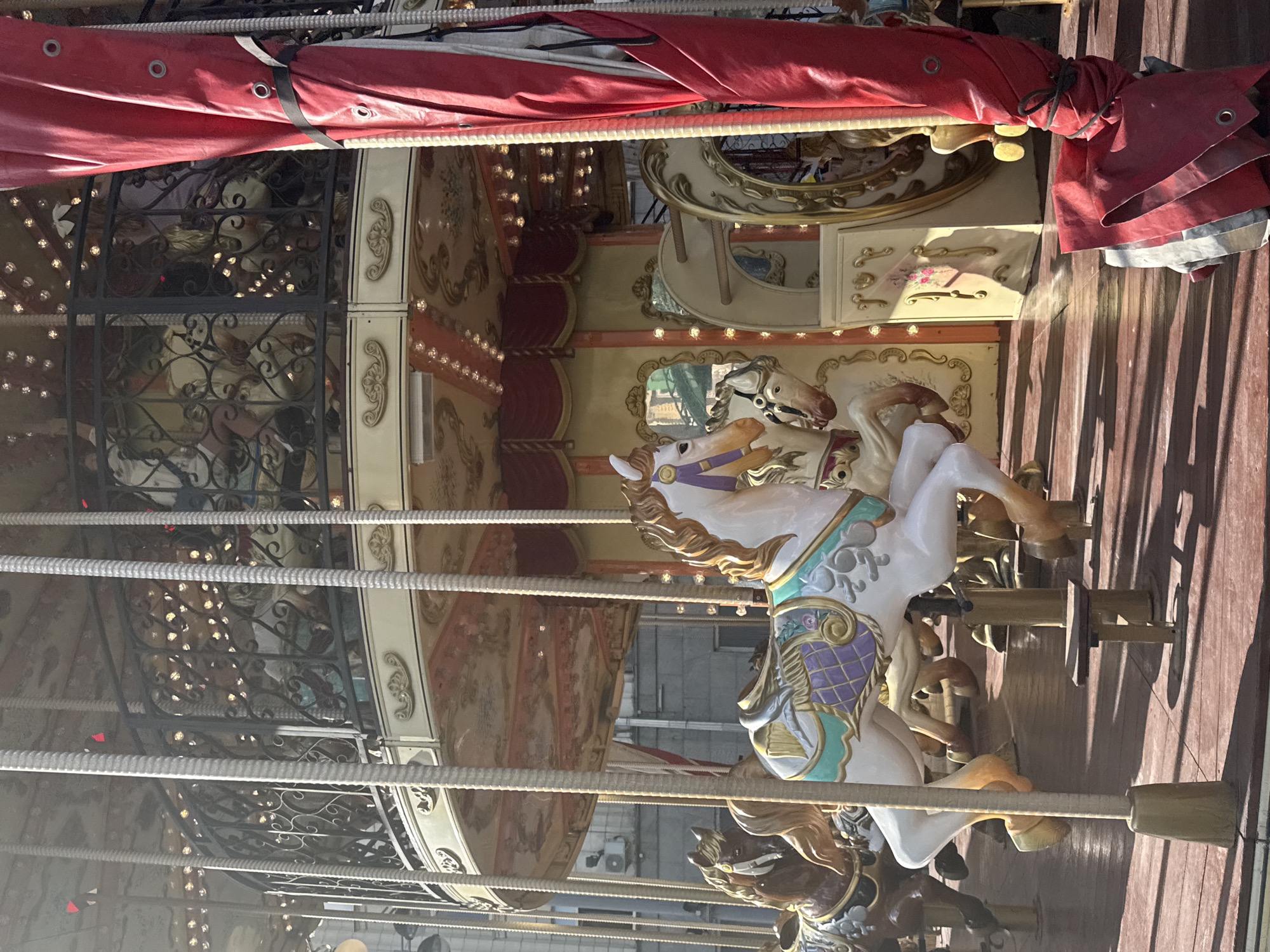 The image depicts a carousel, specifically the "Gatchina" carousel located on Nikolskaya Street in Moscow, Russia. The carousel features several ornate horses, with a prominent white and turquoise horse in the foreground. The horses are attached to poles and positioned on a wooden platform. The carousel's upper levels are visible, adorned with decorative railings and lights. A large red tarp is partially obscuring the right side of the image. The scene is illuminated by sunlight, casting shadows across the wooden floor. The background shows buildings, suggesting an urban setting. No people are visible in the image.