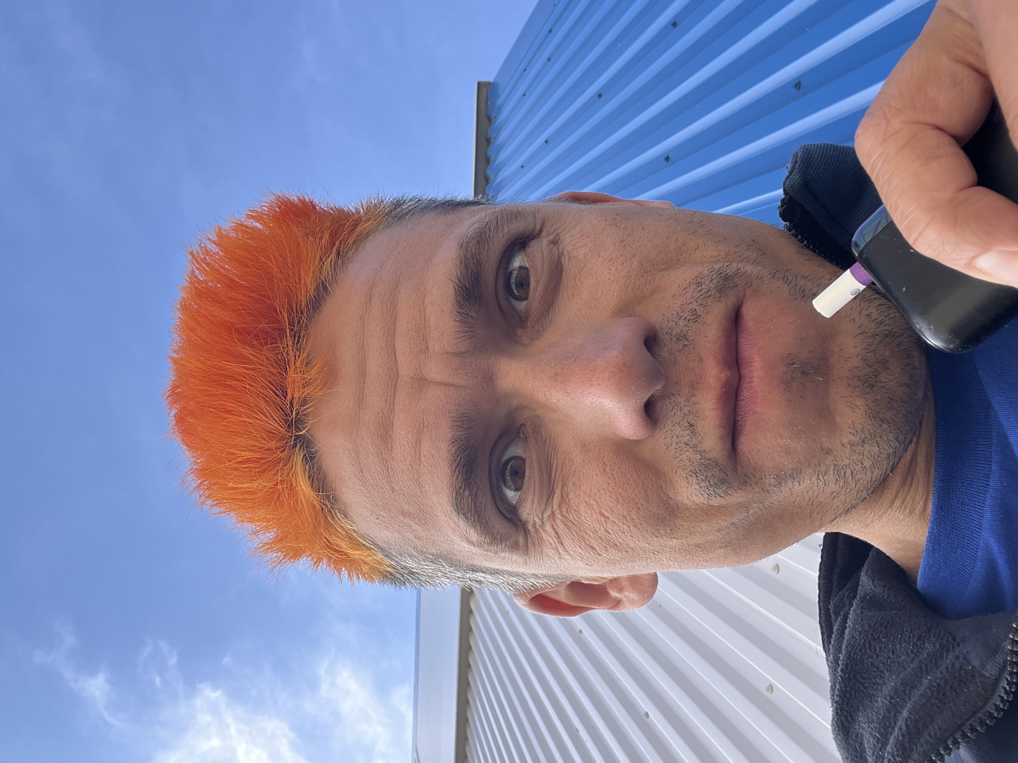 An eye-level portrait captures a man with striking bright orange, spiky hair, partially revealing graying roots. He looks directly at the camera with a neutral, slightly serious expression, showcasing wrinkles on his forehead and stubble on his cheeks and chin. His left eye is clearly visible, with its brown iris.
The man is holding a black and purple electronic device, possibly a vape or electronic inhaler, with a white tip, positioned near his mouth. He is wearing a blue shirt layered under a black zip-up jacket.
The background features a bright blue sky with scattered wispy clouds, indicating clear daytime weather. Below the sky, there are corrugated metal walls; one section is painted blue with horizontal ridges and visible fasteners, while another section, further down, is white with similar construction. These elements suggest an outdoor setting, possibly near an industrial or commercial building in Chaikovsky, Russia. No discernible text is visible in the image.
